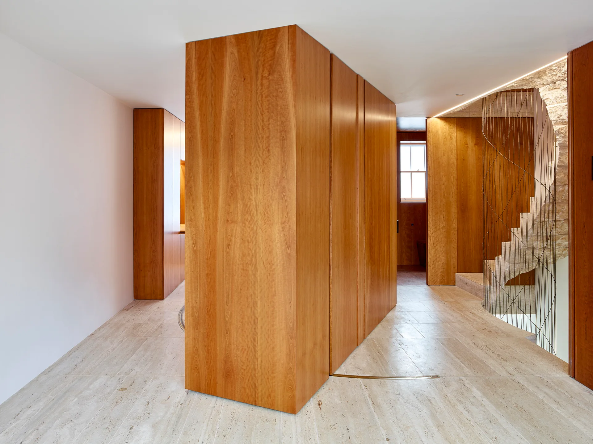 A modern hallway features beige stone tile floors, a large wood-paneled storage column, and a stone spiral staircase with thin vertical railings.