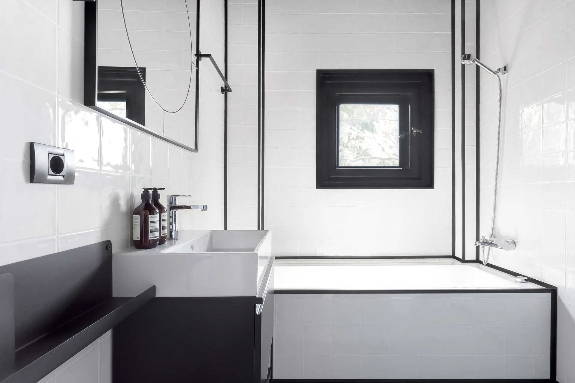 A modern bathroom features glossy white square tiled walls with prominent black vertical accent lines, a black vanity, and a built-in bathtub.