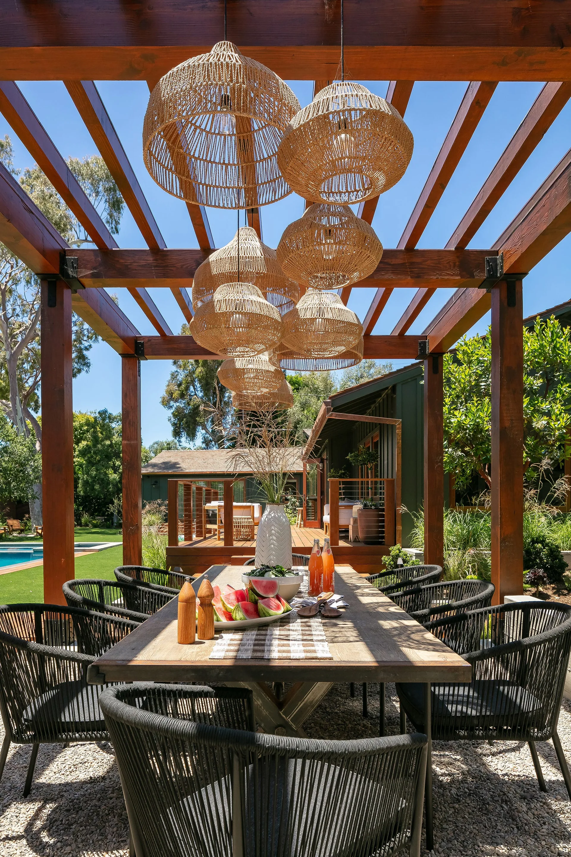 An outdoor wood dining table with black rope chairs sits on gravel under a dark wood pergola hung with several woven basket pendant lights.