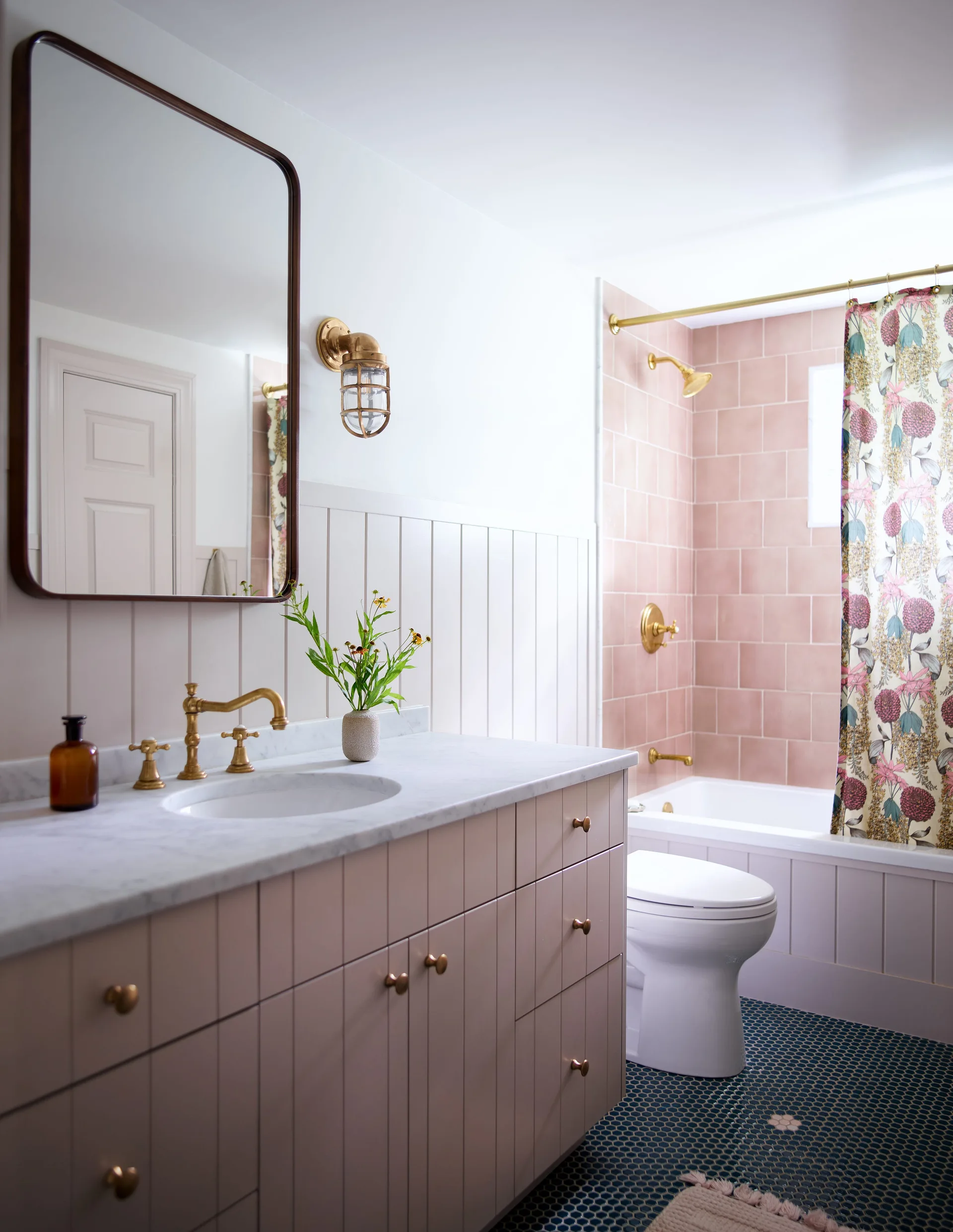 A bathroom with a dusty pink vanity, white stone countertop, brass fixtures, a matching pink tiled tub area, and dark blue hexagonal floor tile.