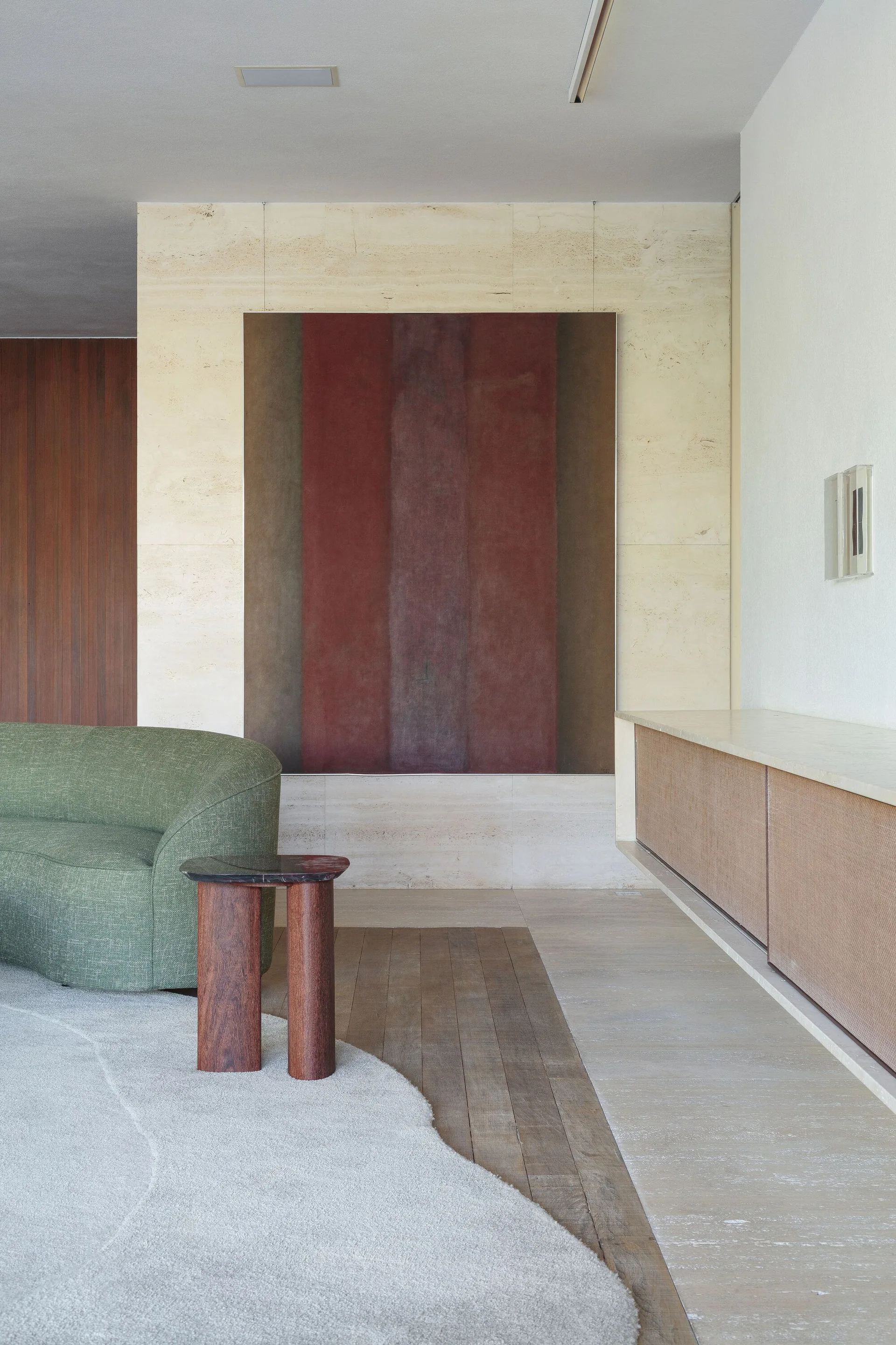 A curved green sofa sits on a textured rug near a travertine stone wall with a large abstract painting and a floating wooden console.