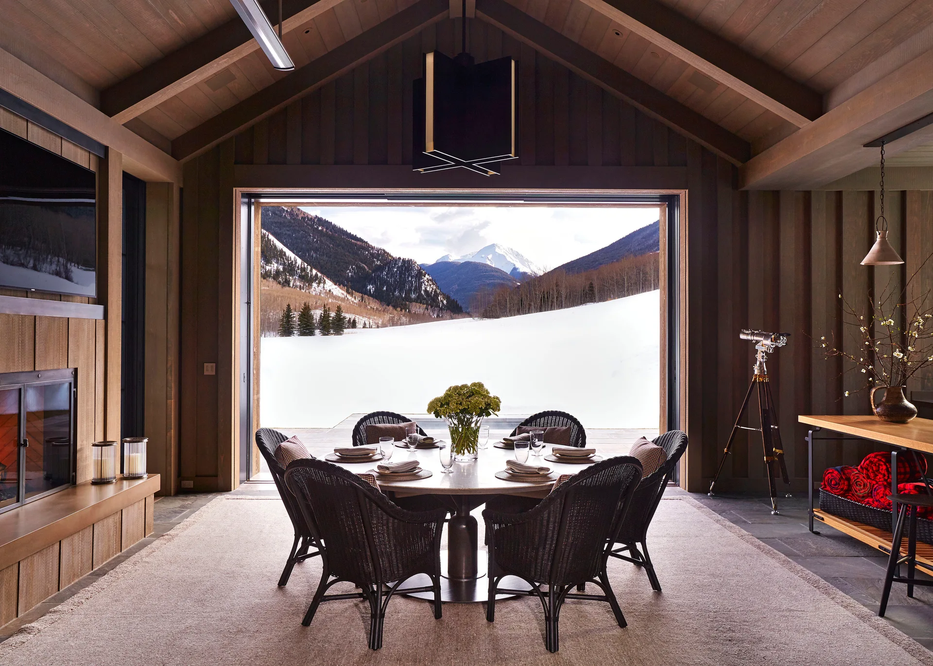 A rustic mountain lodge dining room features a round table with dark woven chairs positioned in front of a wide opening showing a snowy peak.