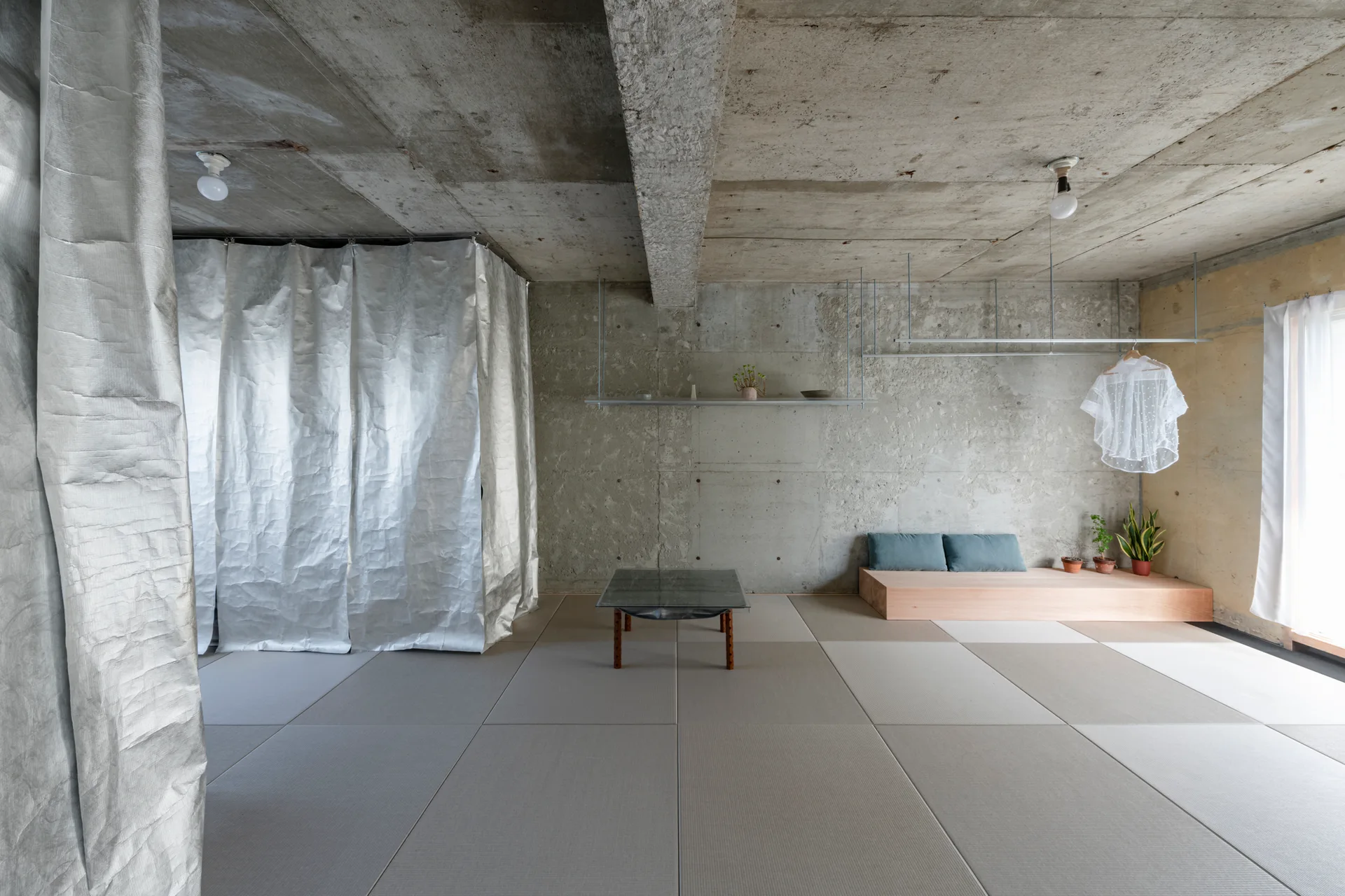 A minimalist room features raw concrete walls and ceiling, modular tatami floor mats, a low wooden platform bench, and a metallic silver curtain.