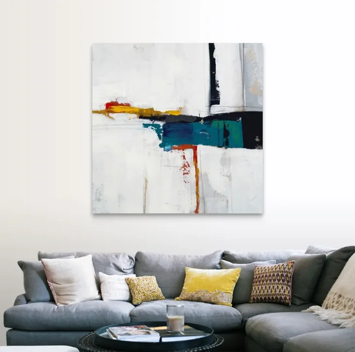 A gray fabric sectional sofa is decorated with patterned yellow throw pillows below a large square abstract painting on a white wall.