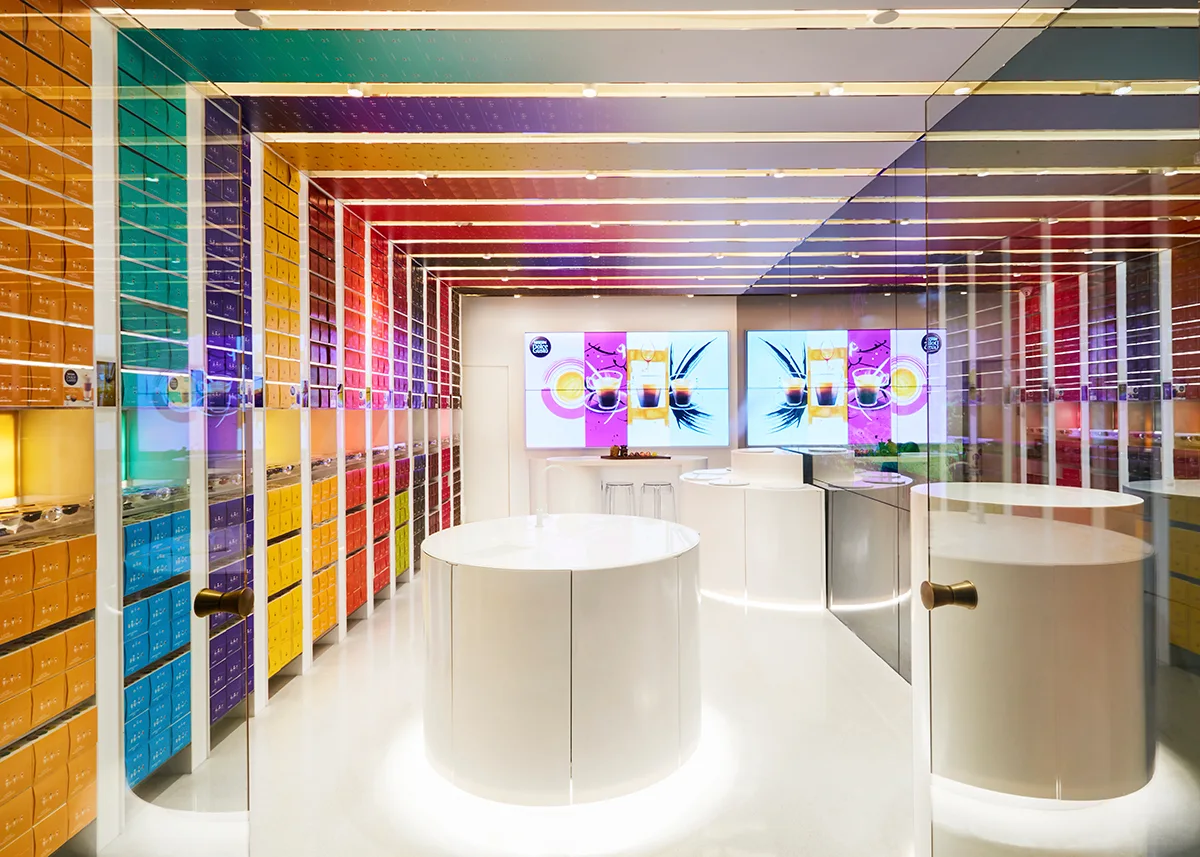 A bright retail showroom features rainbow colored product shelving along the walls and glossy white circular display counters on a reflective floor.
