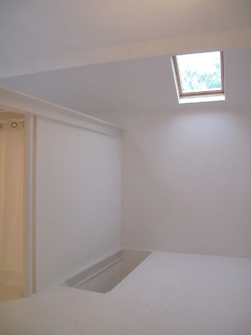 A minimalist all-white room features a rectangular ceiling skylight, a recessed floor opening, and a partial partition wall with a white curtain.