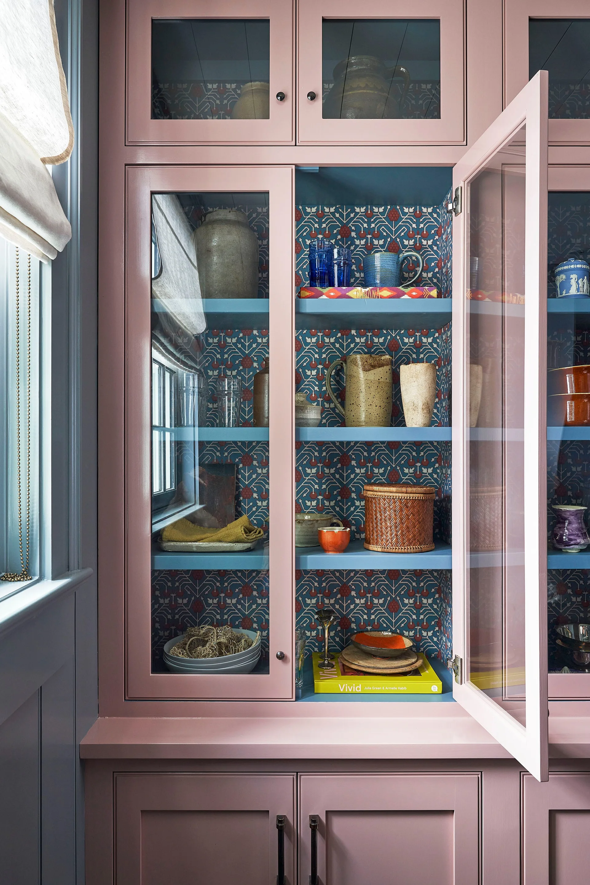 A dusty pink wooden display cabinet with glass doors features blue shelves lined with botanical wallpaper and filled with ceramics.