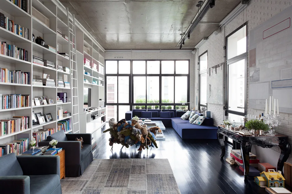 An industrial loft living room with floor-to-ceiling bookshelves, a library ladder, a blue sectional sofa, and an exposed concrete ceiling.