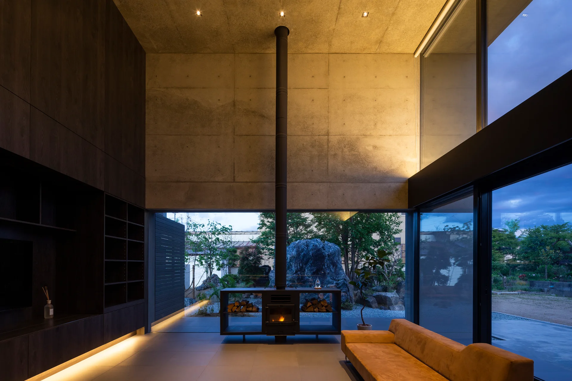 A minimalist living room with exposed concrete walls, a central black metal fireplace, custom dark wood cabinetry, and large windows opening to a garden.