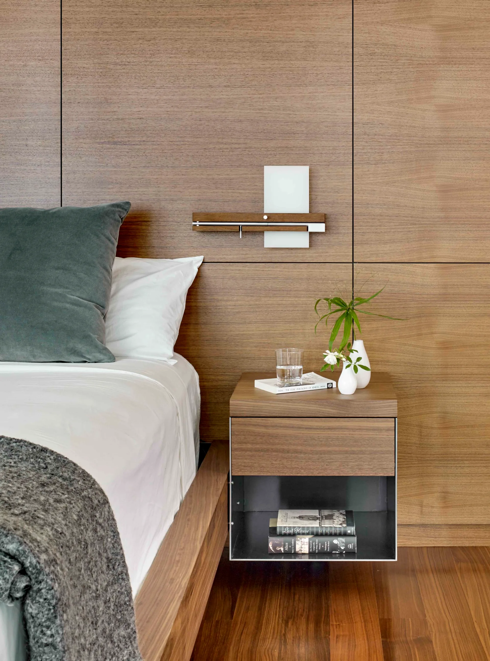 A floating wooden nightstand with books and a vase is mounted against a matching wood-paneled bedroom wall next to a bed.