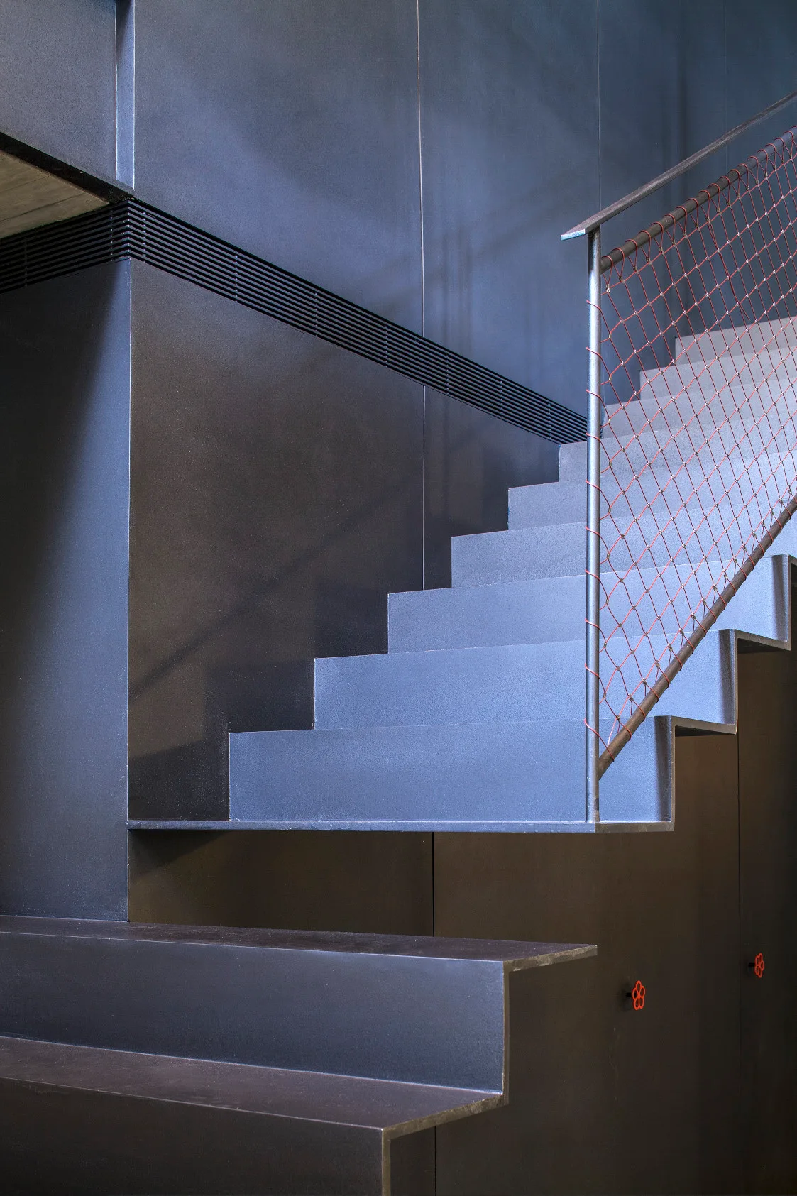 An industrial charcoal metal staircase with angular steps features a thin metal railing filled with red wire mesh netting against dark paneled walls.