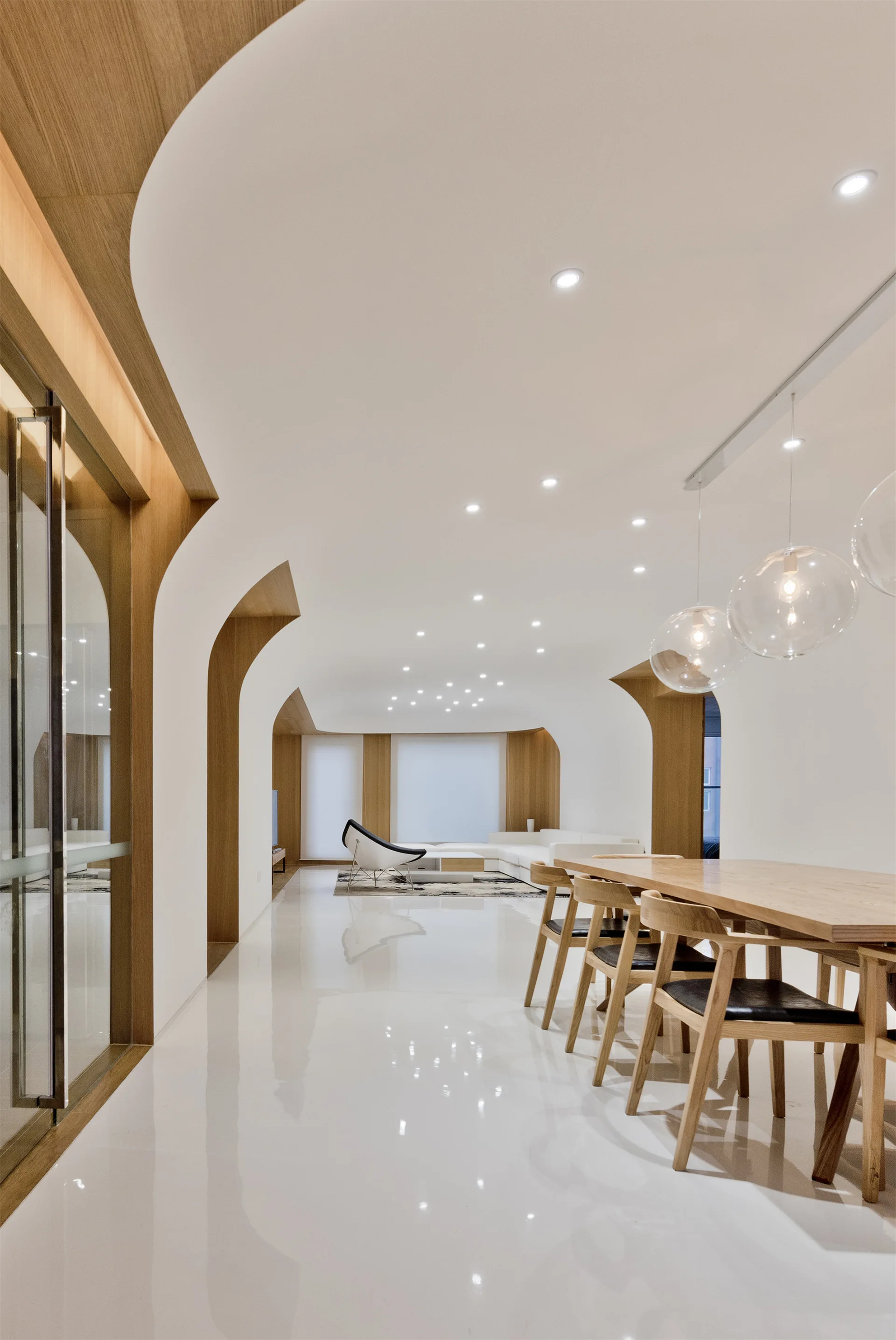 A minimalist dining room features a wood dining table, spherical glass pendant lights, glossy white floors, and curved wood-trimmed archways.