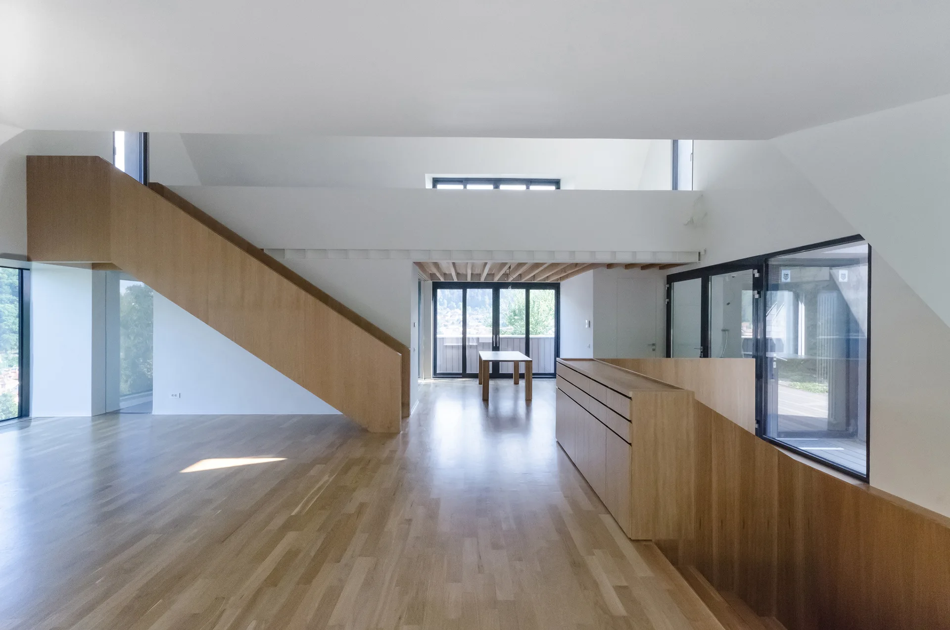 A spacious minimalist interior features light hardwood floors, white walls, and prominent wood-clad staircases leading to an upper loft level.