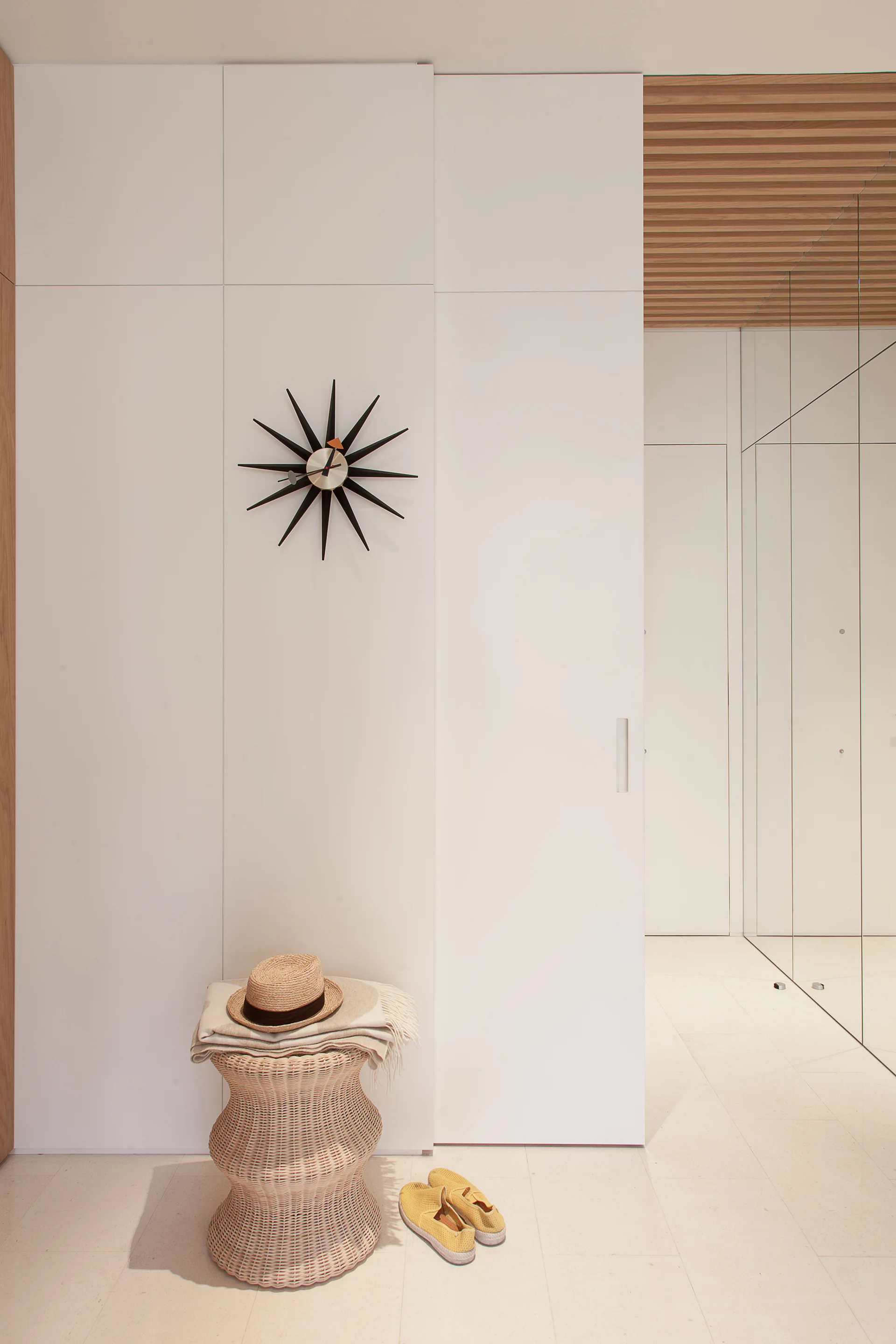 A minimalist white entryway features floor-to-ceiling built-in closets, a black starburst clock, a textured wicker stool, and a slatted wood ceiling.