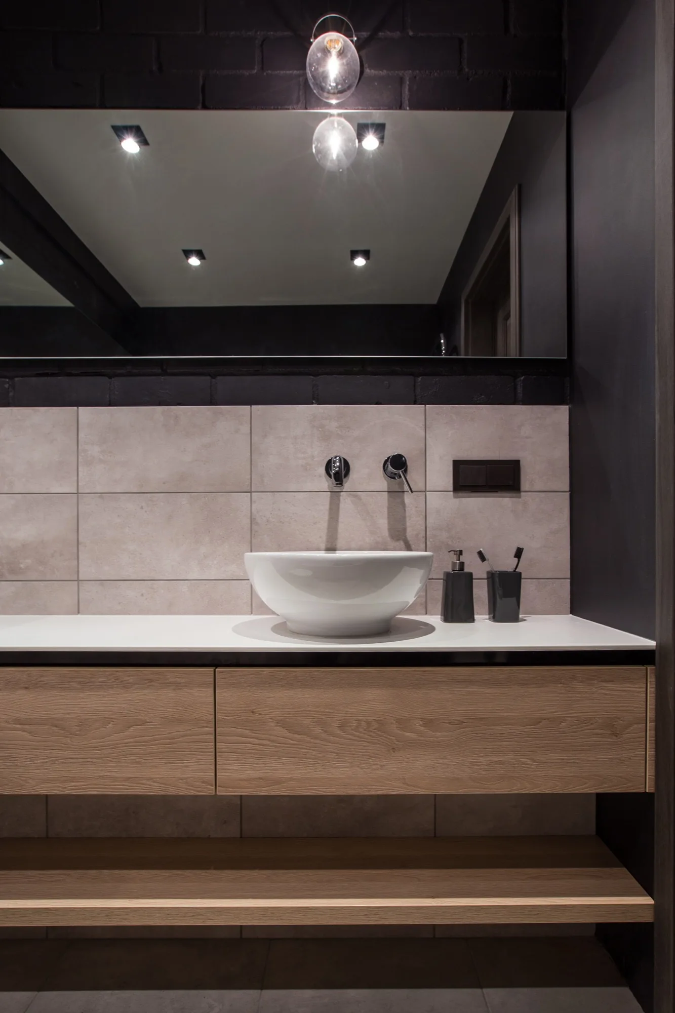 A bathroom vanity features a white vessel sink, a floating light wood cabinet, grey tiled backsplash, and a large mirror beneath a black brick wall.