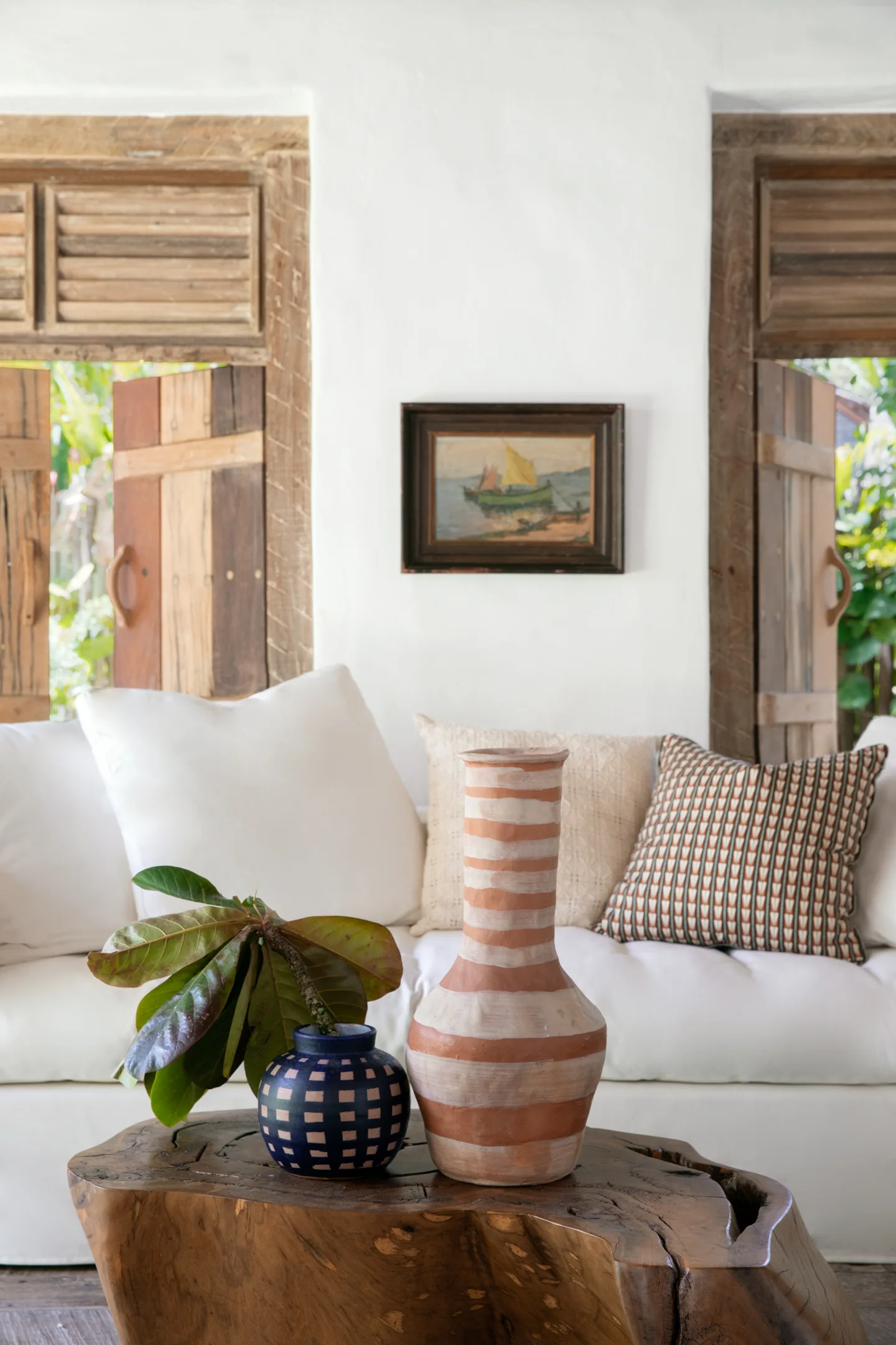 A white sofa with patterned pillows sits behind a rustic wood coffee table styled with ceramic vases against a plaster wall with wooden shutters.