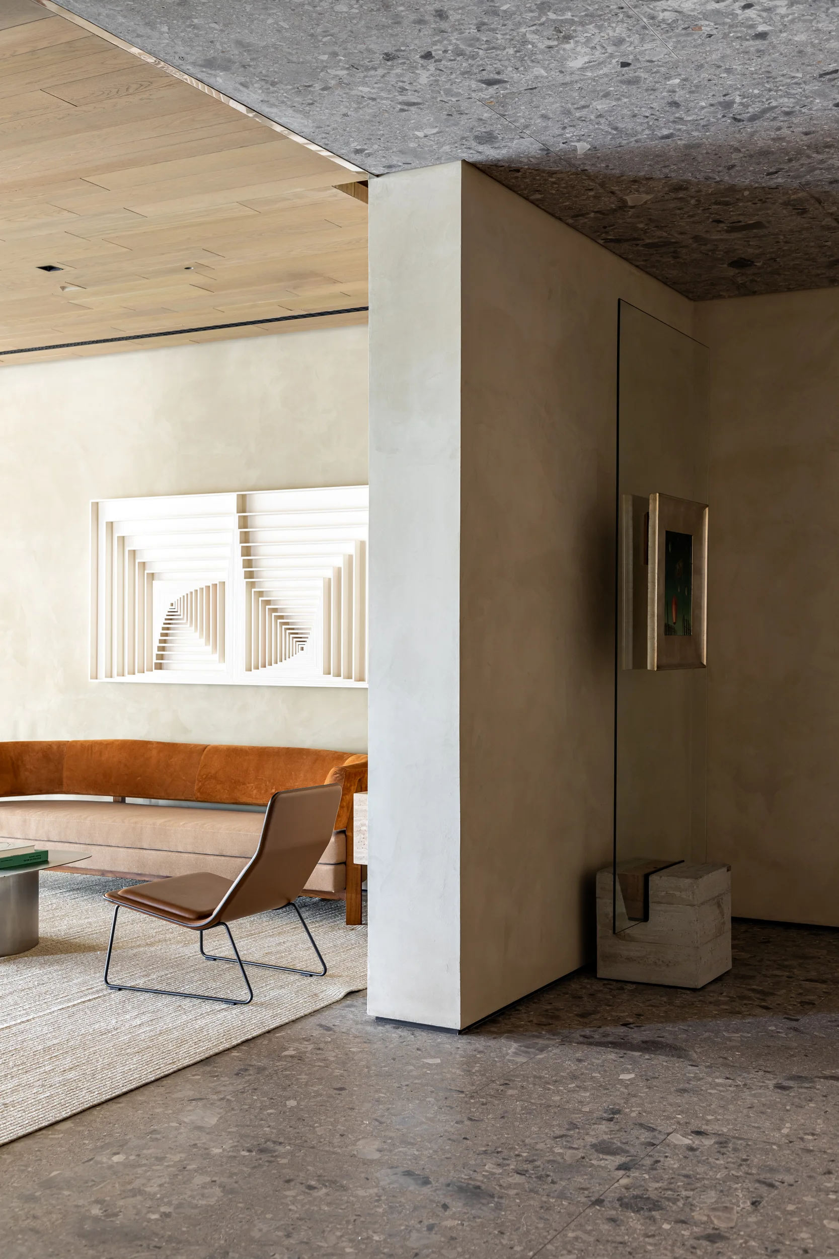 A minimalist living room with a curved terracotta orange sofa, brown leather chair, textured rug, plaster walls, and a white geometric relief sculpture.