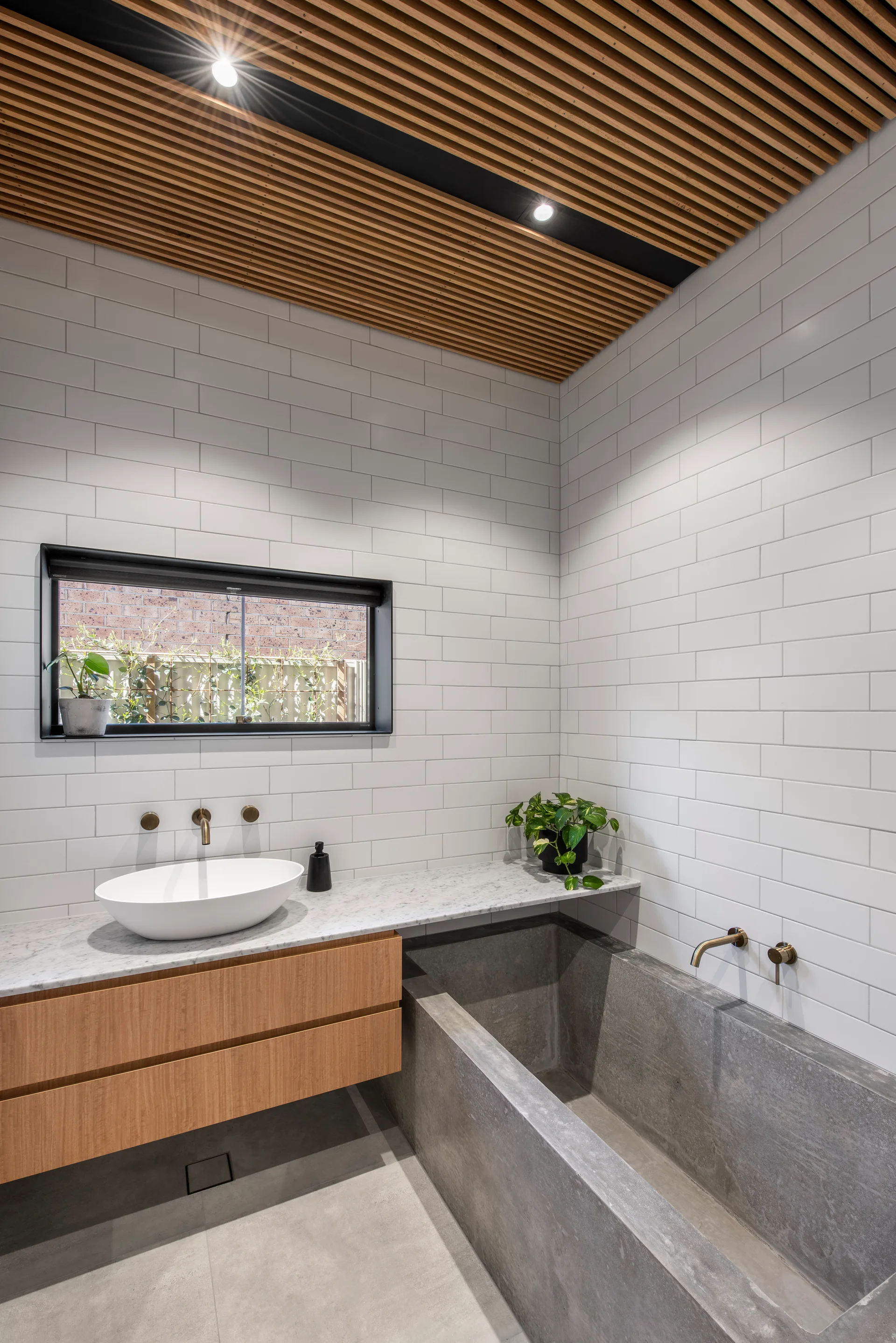 A modern bathroom features a rectangular concrete bathtub, a wooden floating vanity with a marble top, white subway tiles, and a slatted timber ceiling.