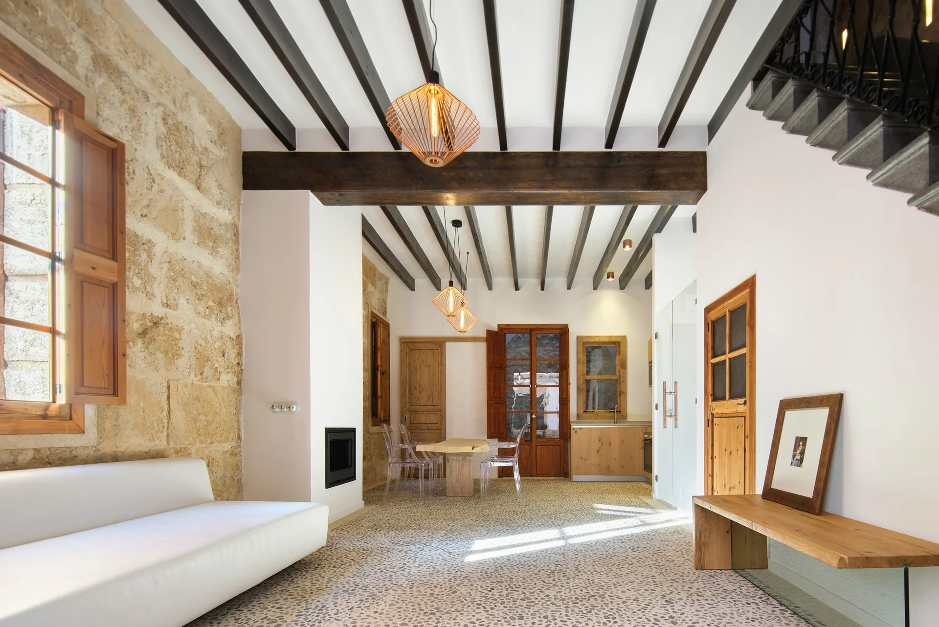 A rustic modern living area features an exposed stone wall, dark wooden ceiling beams, a pebble-stone floor, and a white low-profile sofa.