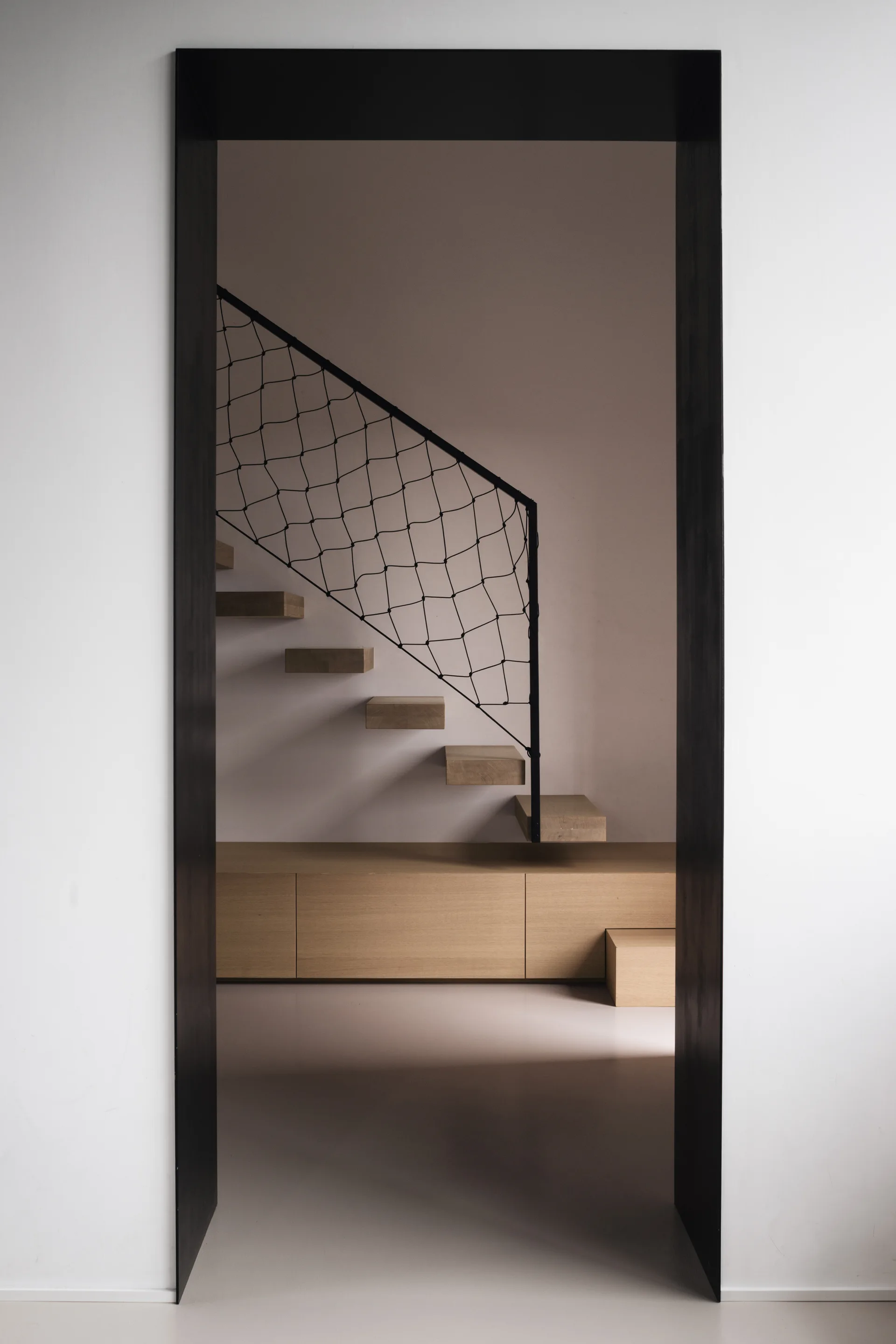 A dark framed doorway looks in on a minimalist staircase with floating wooden treads, a black mesh railing, and a matching low wooden storage cabinet.