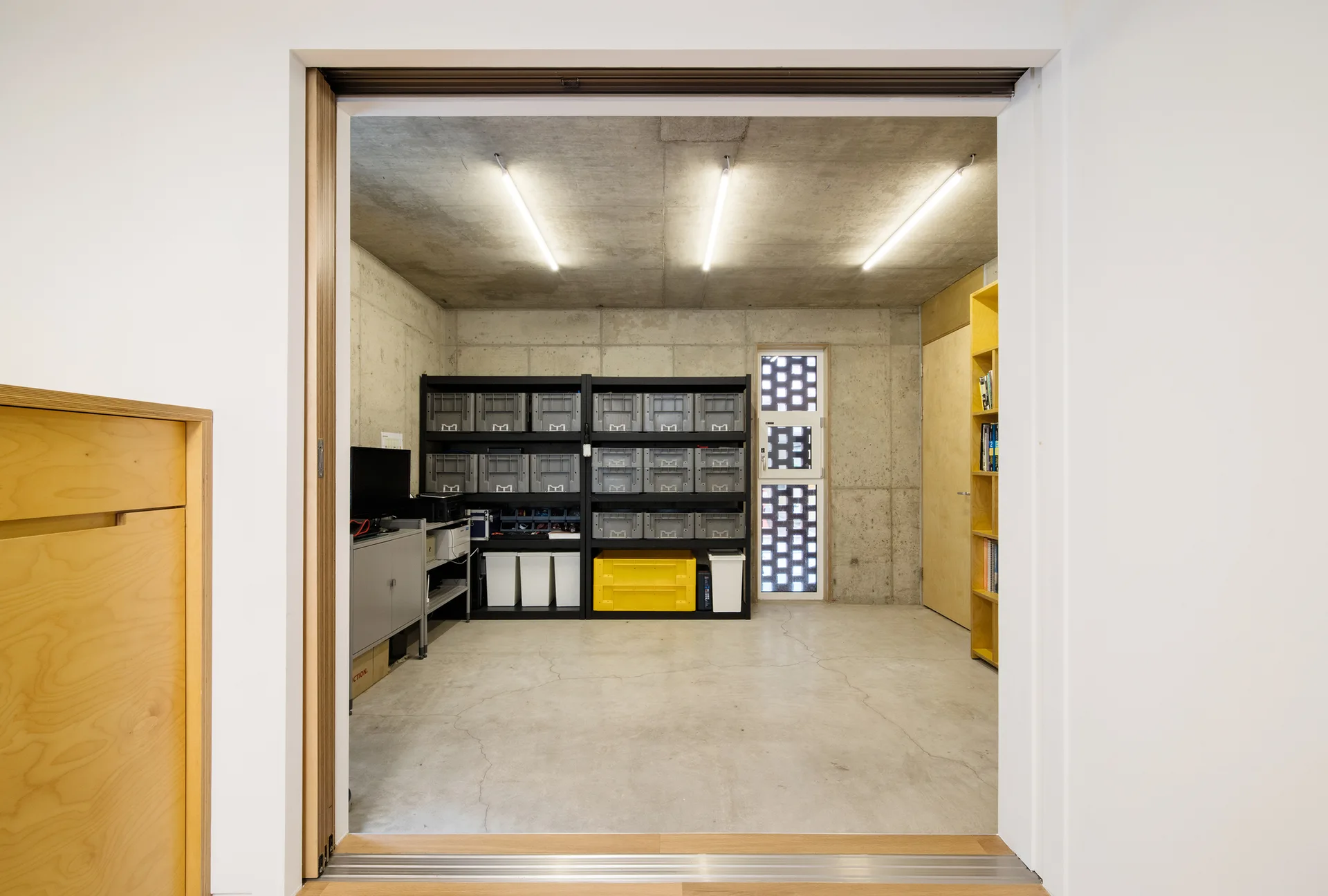 A storage room viewed through a doorway, featuring exposed concrete walls, industrial black shelving with grey bins, and a patterned concrete block window.