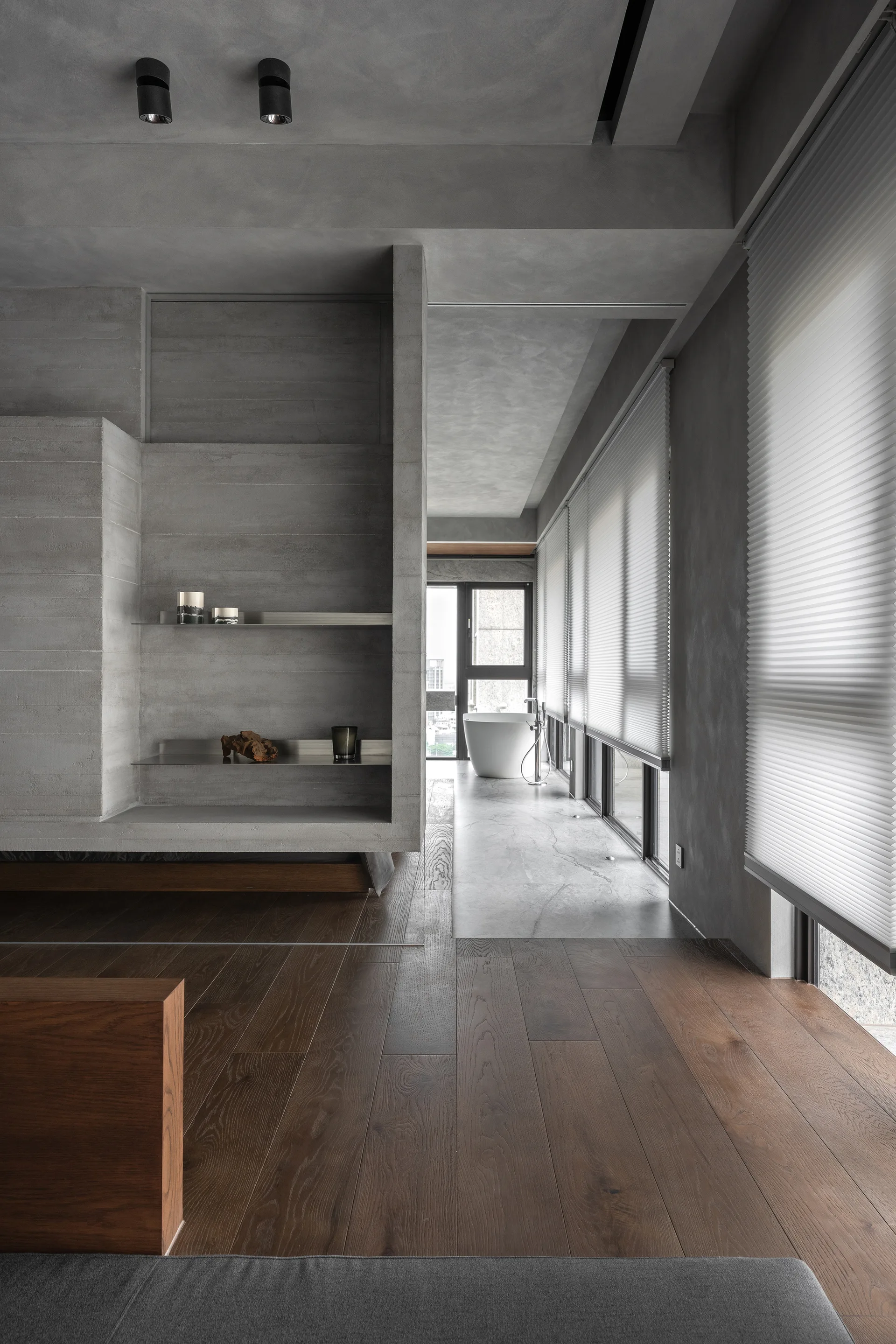 A modern concrete bathroom with a freestanding white tub, dark wood flooring, pleated window blinds, and a grey concrete niche with metal shelving.
