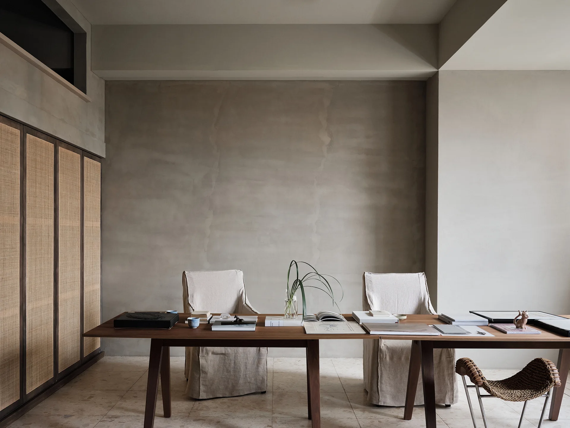 A long wooden desk sits in front of a textured plaster wall, paired with linen-slipcovered chairs and a dark wood cane cabinet.