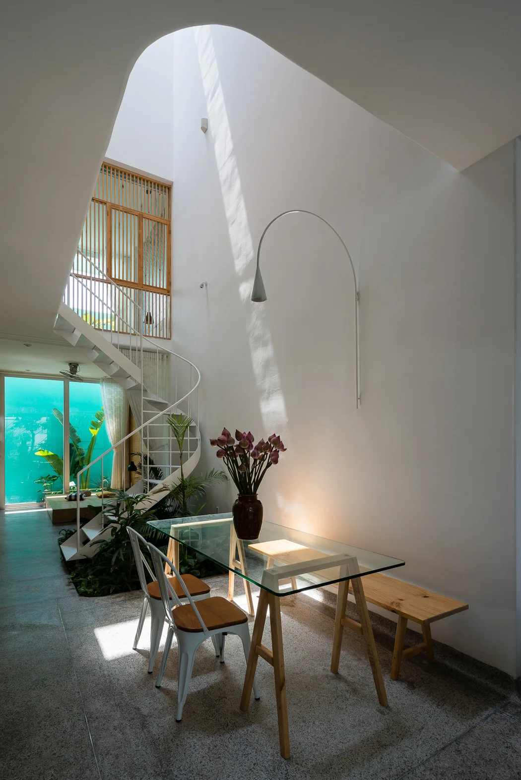A modern dining area with a glass table beneath a high white atrium lightwell featuring a spiral staircase and indoor plants.