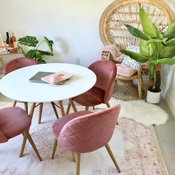 A round white dining table is surrounded by four pink velvet chairs on a patterned pink rug next to a wicker peacock chair.