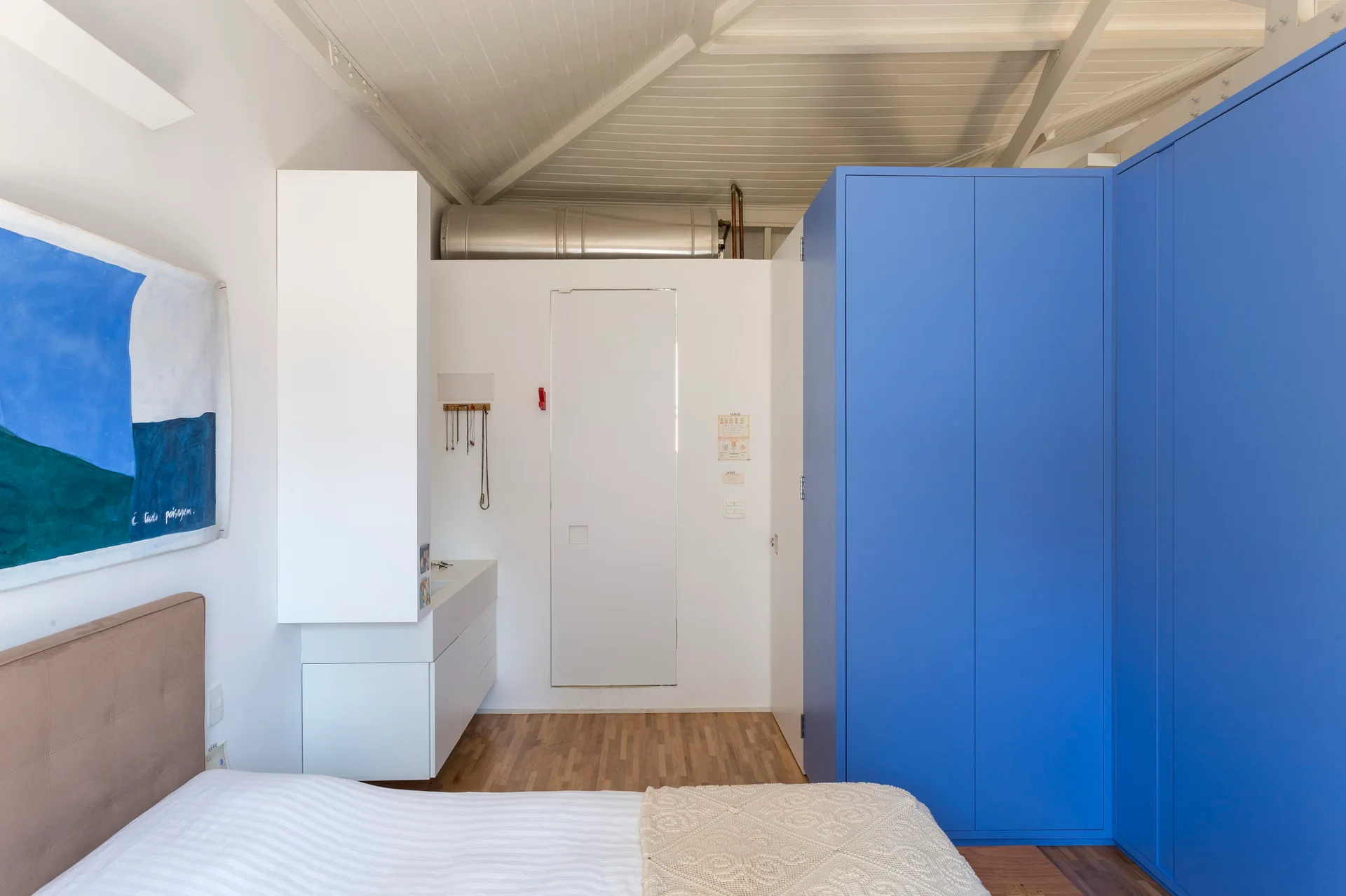 A modern loft bedroom features a bold matte blue corner wardrobe, white walls, and a sloped ceiling with exposed white wooden beams.