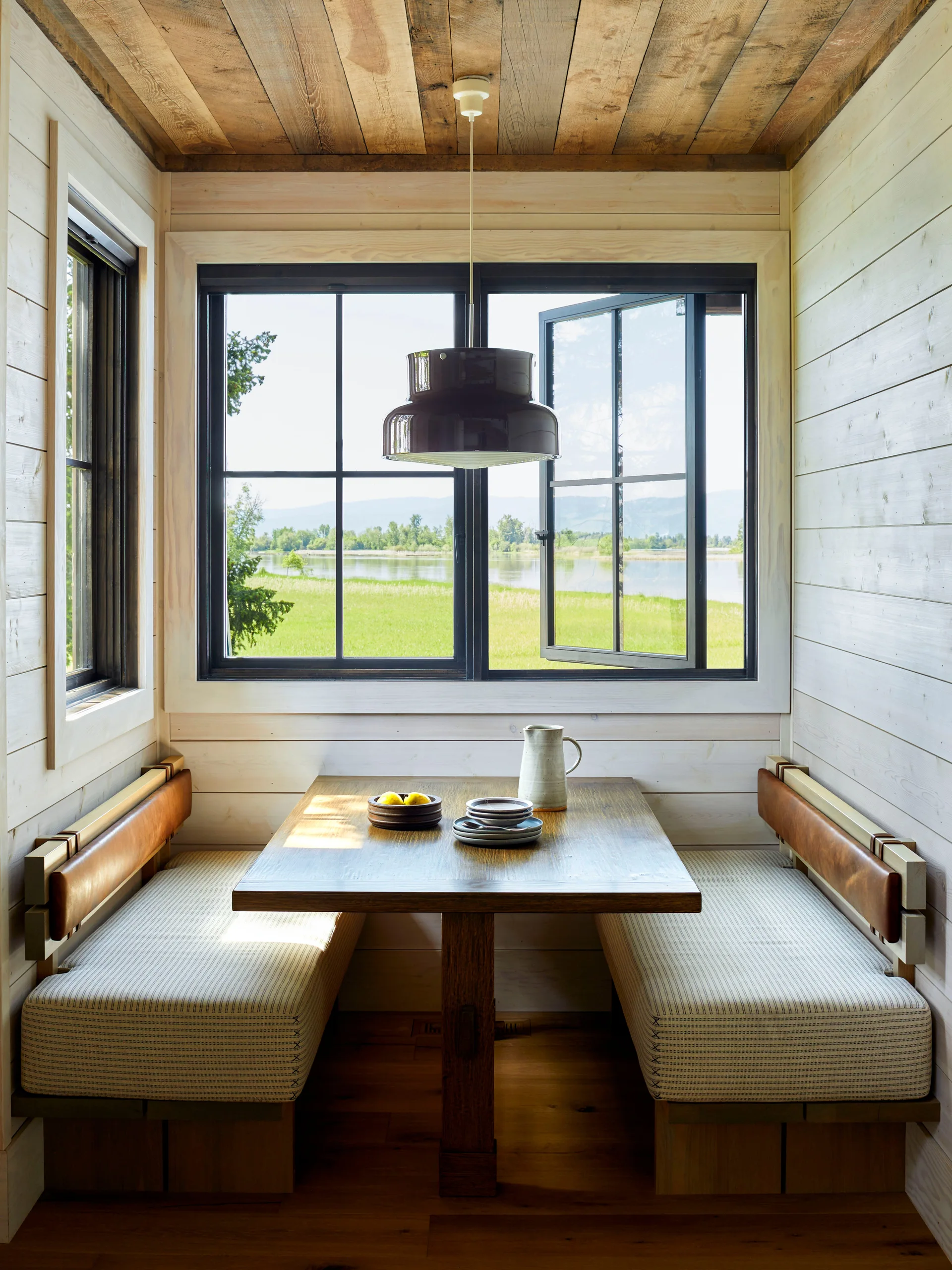 A rustic breakfast nook features built-in benches with striped cushions, a wooden table, and a window displaying a view of water and grassy hills.
