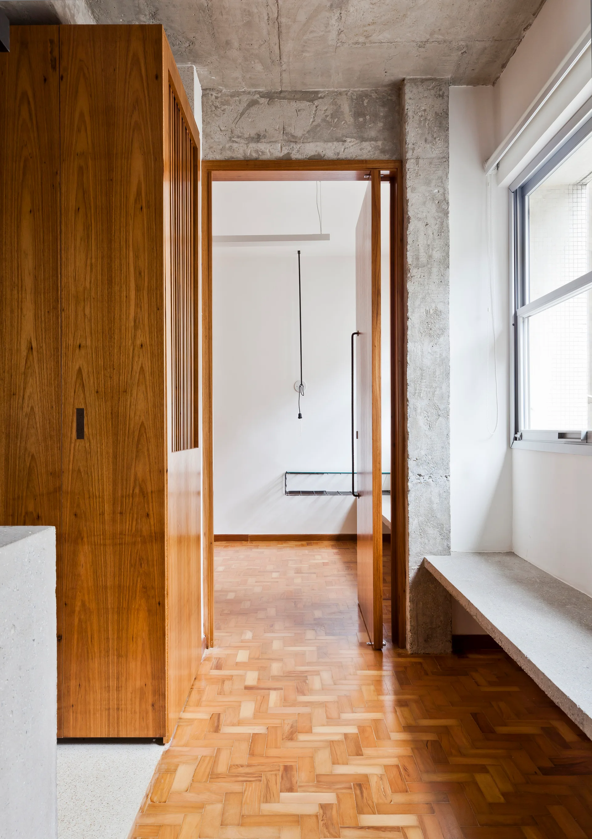 A minimalist hallway featuring warm wood parquet flooring, a tall wooden cabinet, exposed concrete structural beams, and a concrete bench under a window.