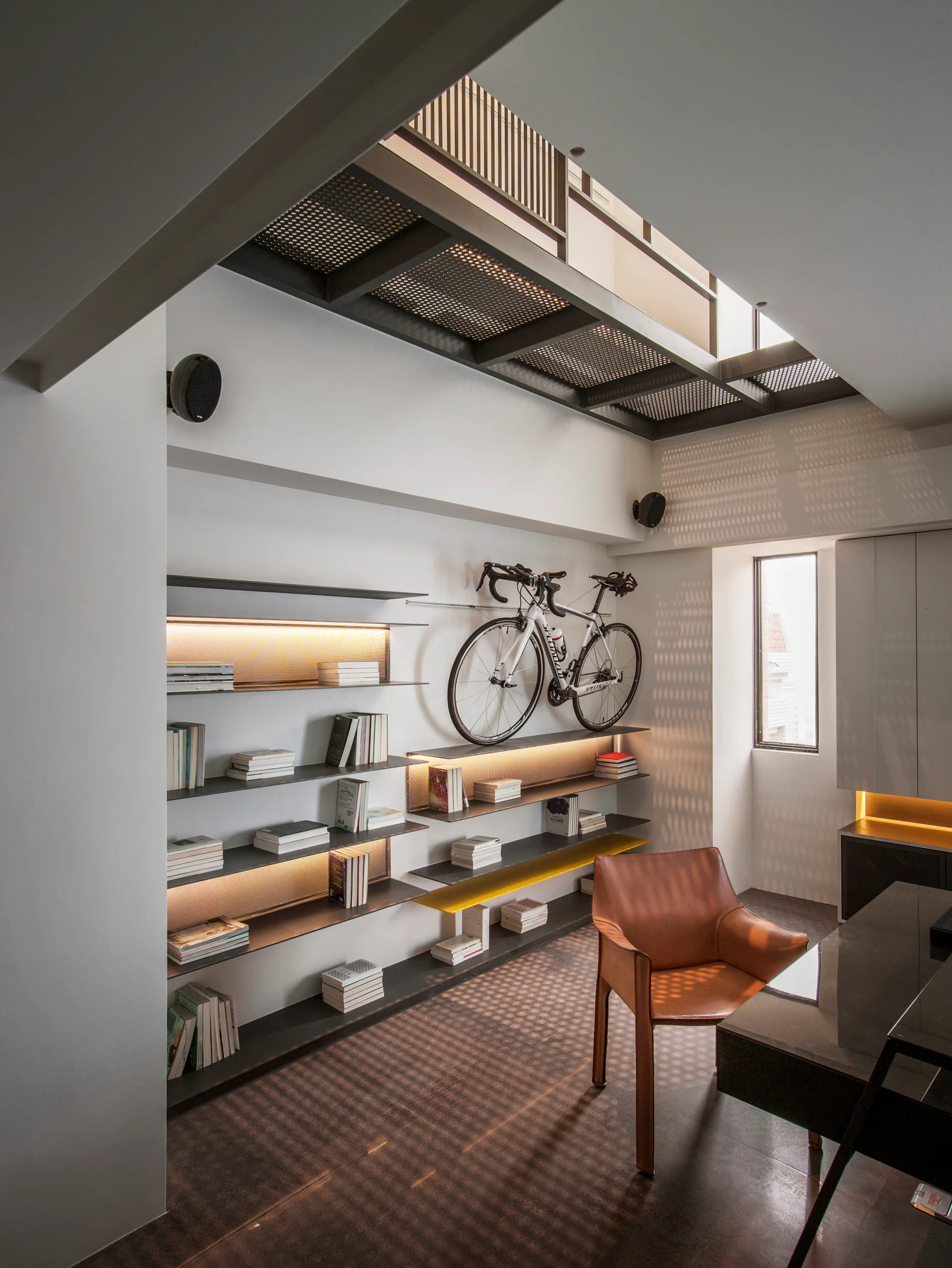 A modern home office features wall-mounted metal shelves with integrated lighting, a mounted bicycle, and a leather armchair.