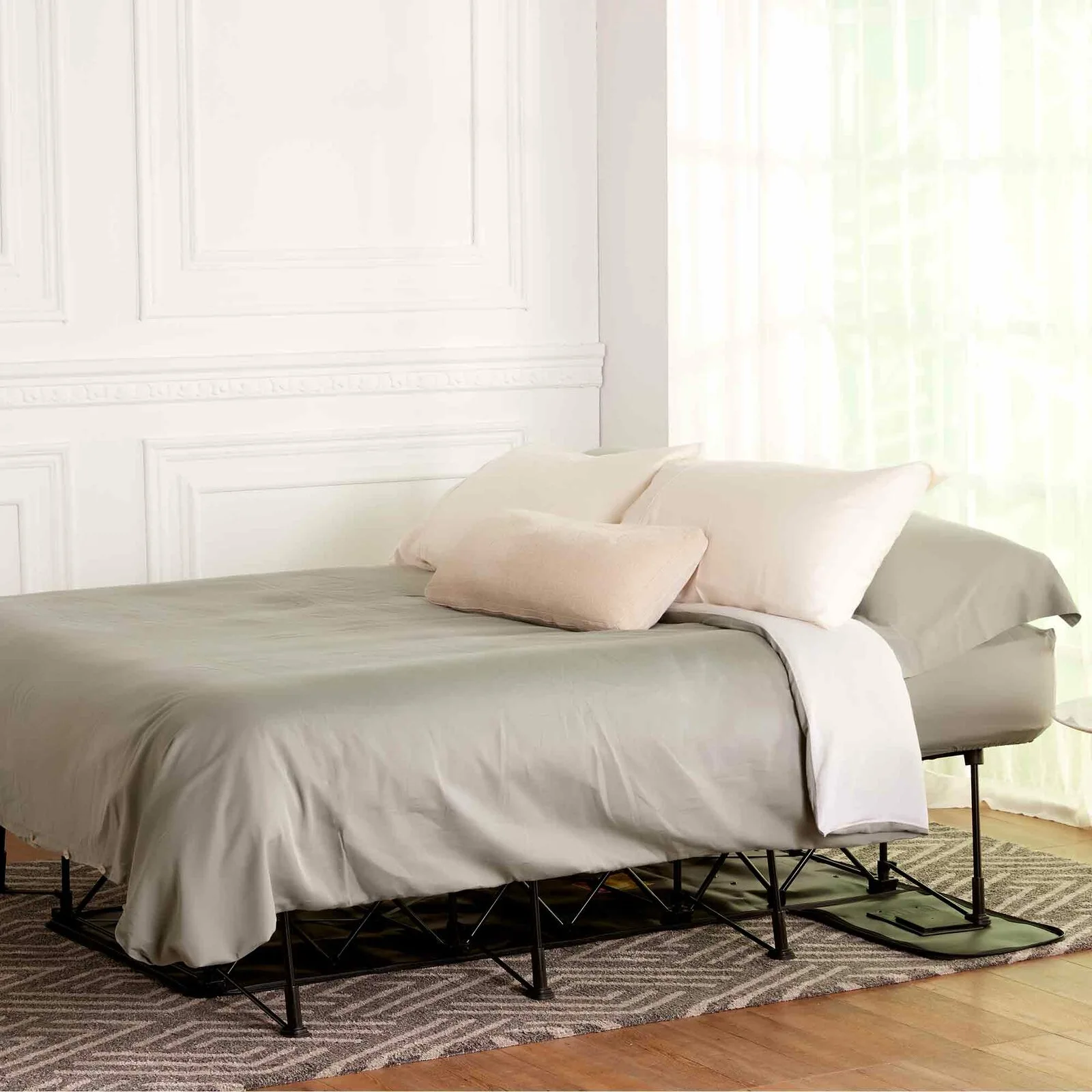 A portable folding bed with grey linens and pillows sits on a geometric patterned rug against a white paneled wall.