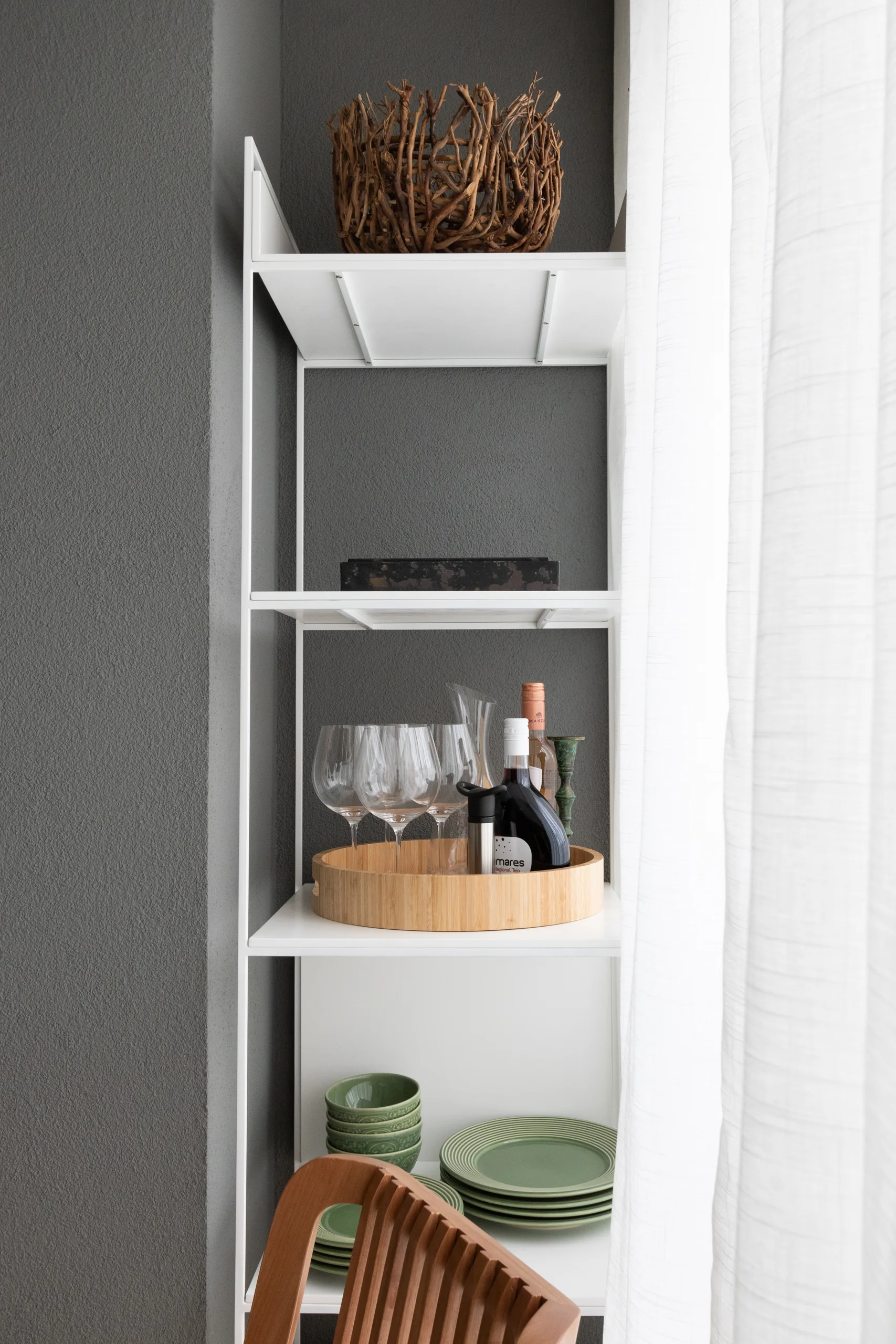 A white metal open shelving unit styled with green ceramic plates, glassware on a wooden tray, and a branch basket against a dark grey textured wall.