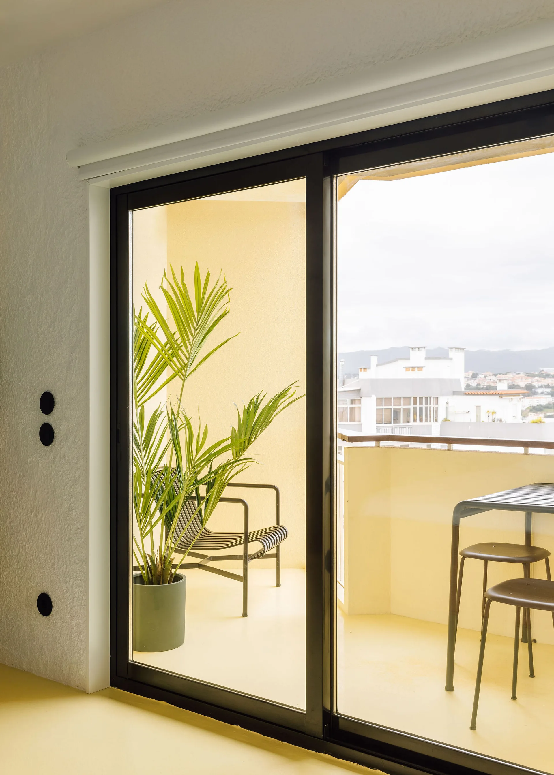 A view through a black-framed sliding glass door reveals a yellow painted balcony with a striped armchair, a small table, and a potted plant.