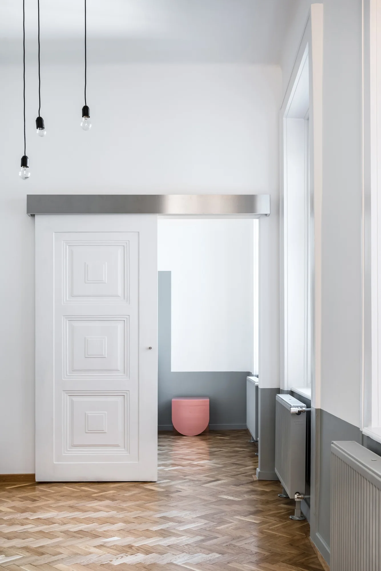 A bright hallway features a white paneled sliding door, herringbone wood floors, grey radiators, and suspended bare bulb pendant lights.