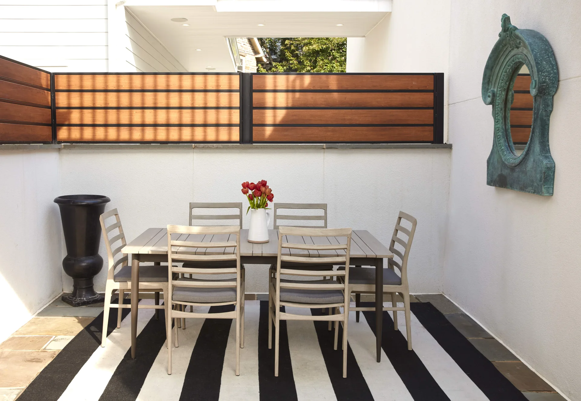 A modern patio dining table and six chairs sit on a black and white striped rug next to a white stucco wall and horizontal wood fence.