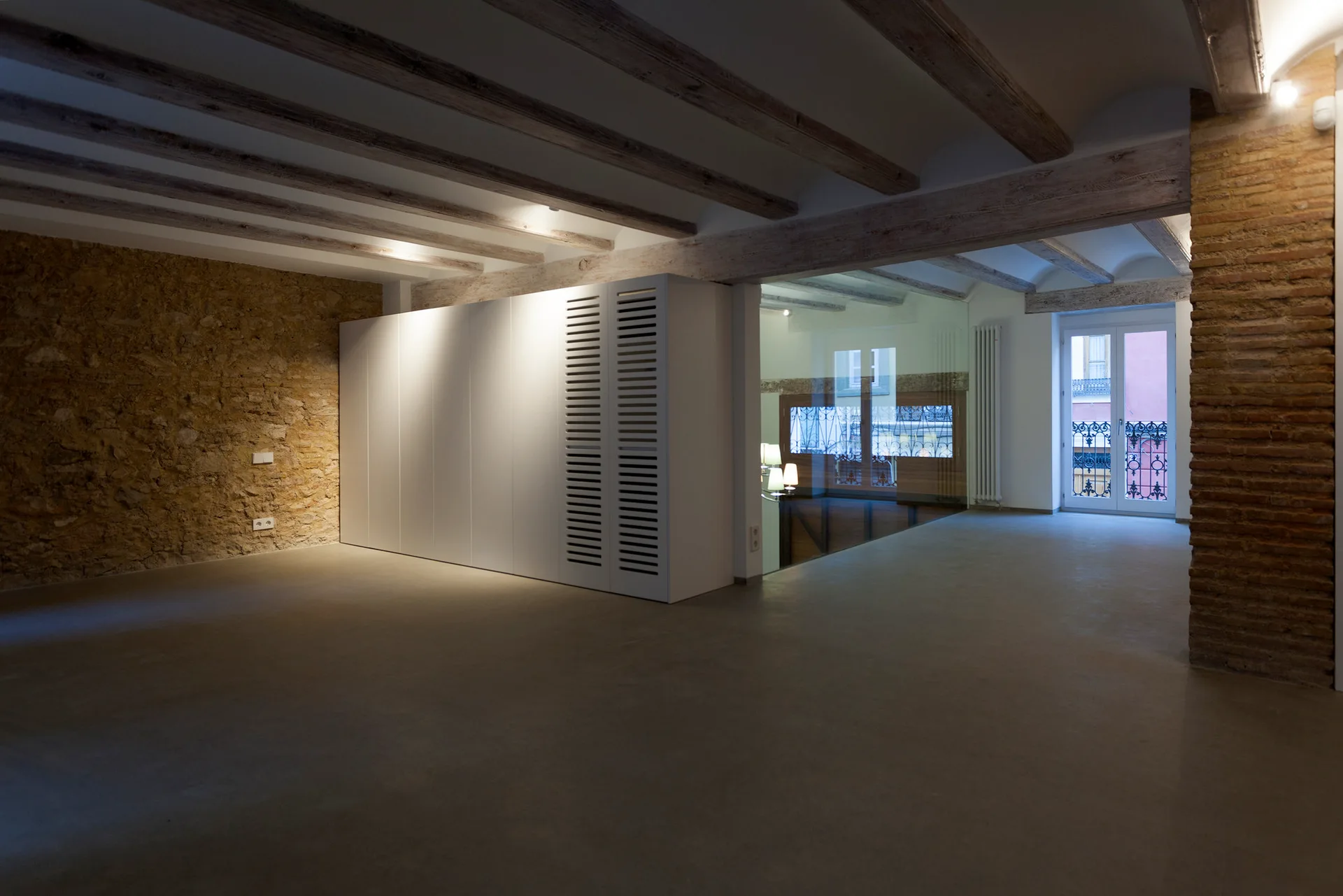 An open room features exposed wood ceiling beams, a rough stone wall, a modern white partition wardrobe, and smooth grey concrete floors.