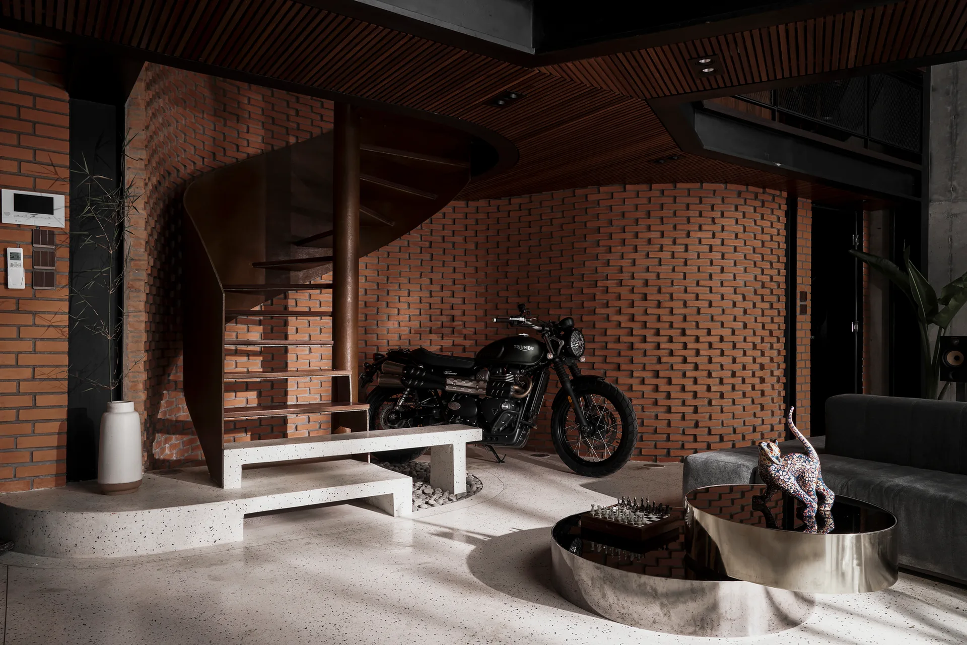 An industrial living room featuring a metal spiral staircase against a curved red brick wall with a parked motorcycle and terrazzo floor.