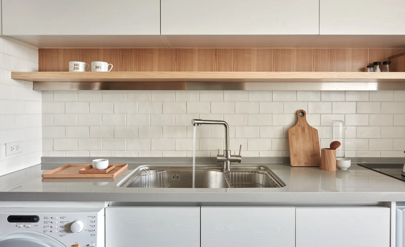 A modern kitchen sink with a brushed metal faucet, a grey stone countertop, a white subway tile backsplash, and a wooden floating shelf.