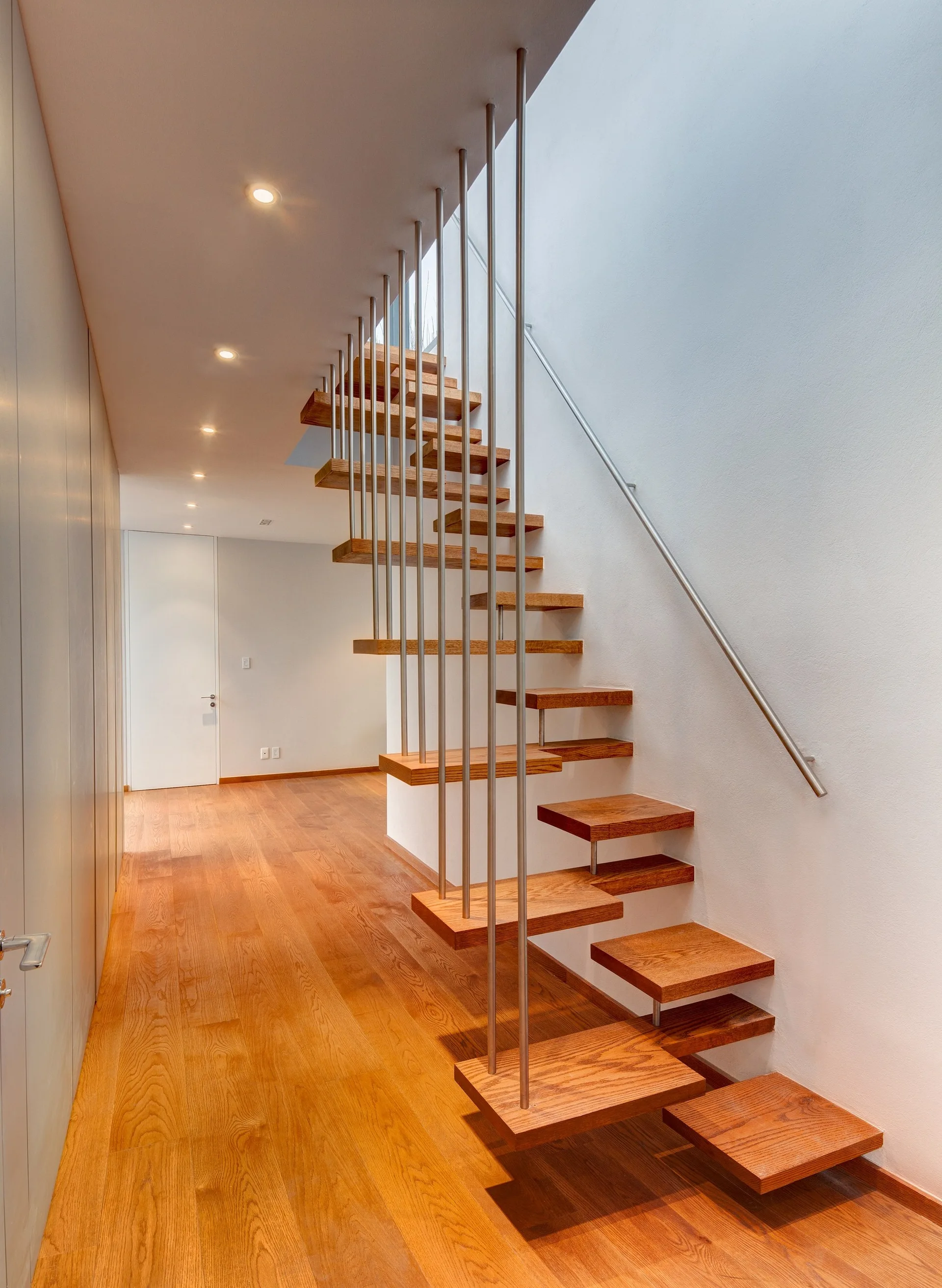 A modern floating wooden staircase with thin vertical metal rods and matching wood floors in a minimalist white hallway with recessed lighting.