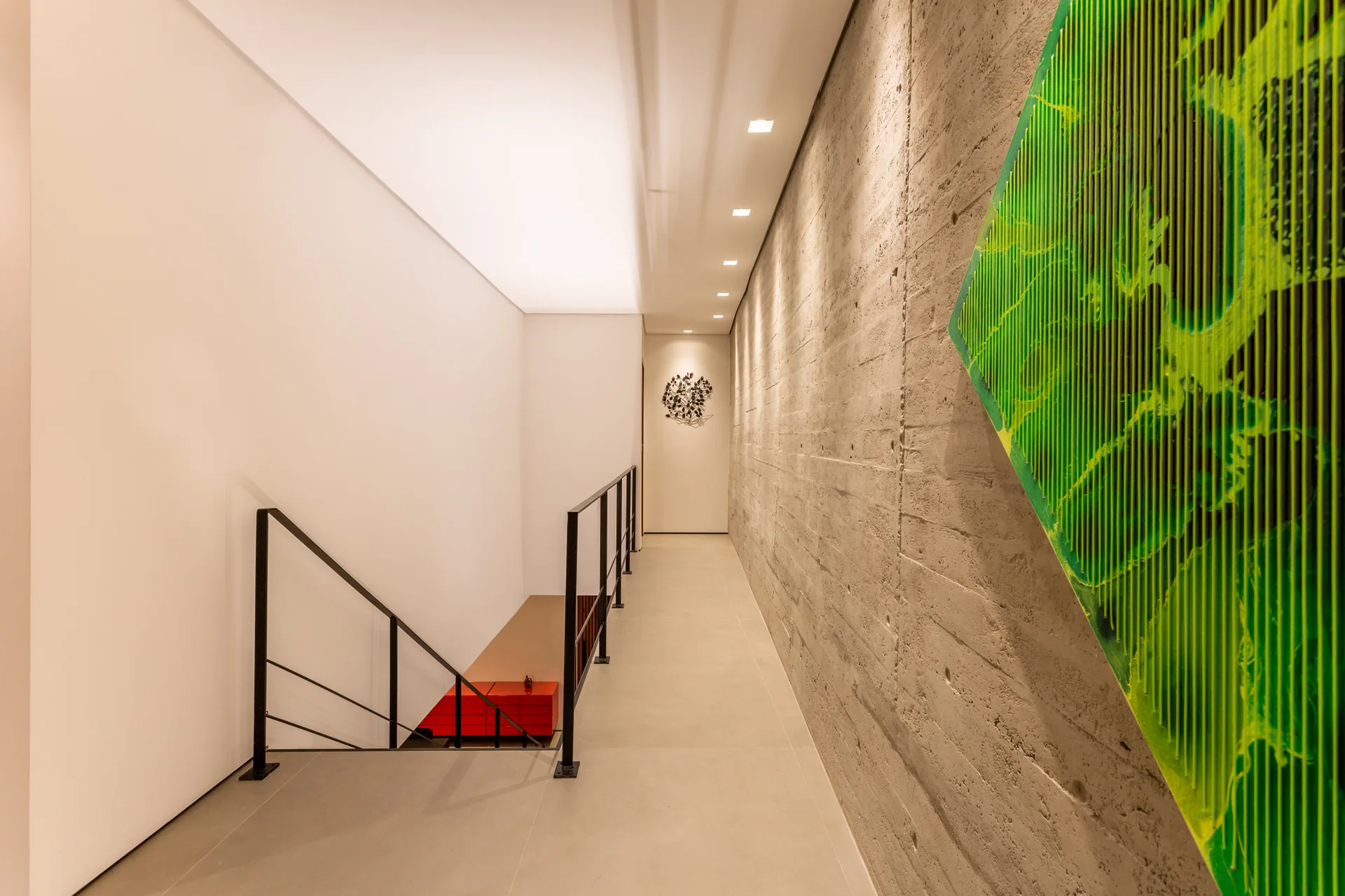 A modern hallway features a textured board-formed concrete wall with green art, contrasted by a white plaster wall and a black metal stair railing.