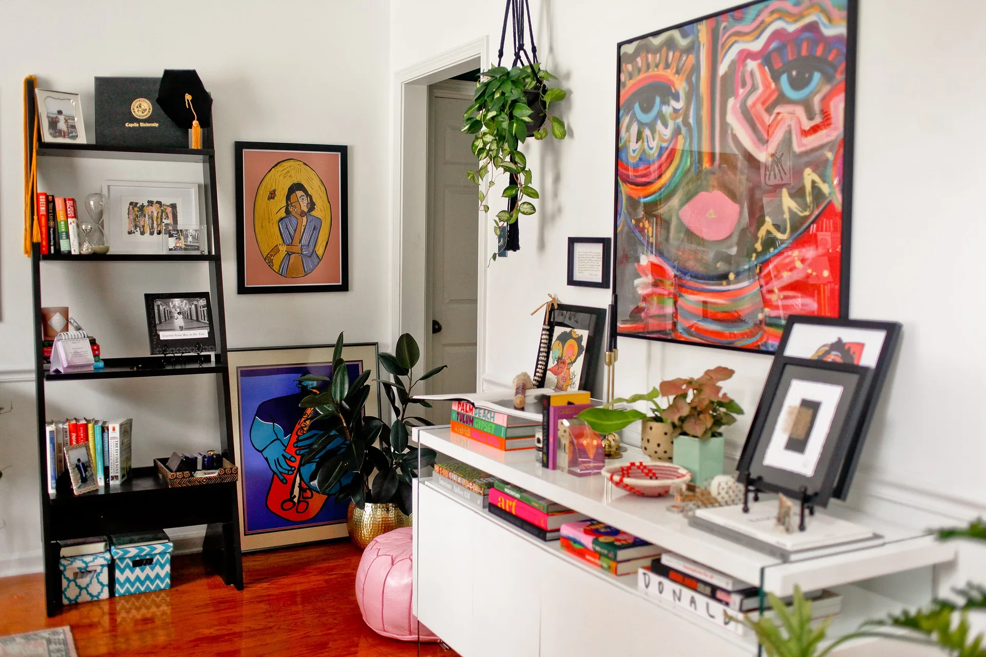 An eclectic room features a white console table stacked with books, a black ladder shelf, and vibrant framed art on a white wall.