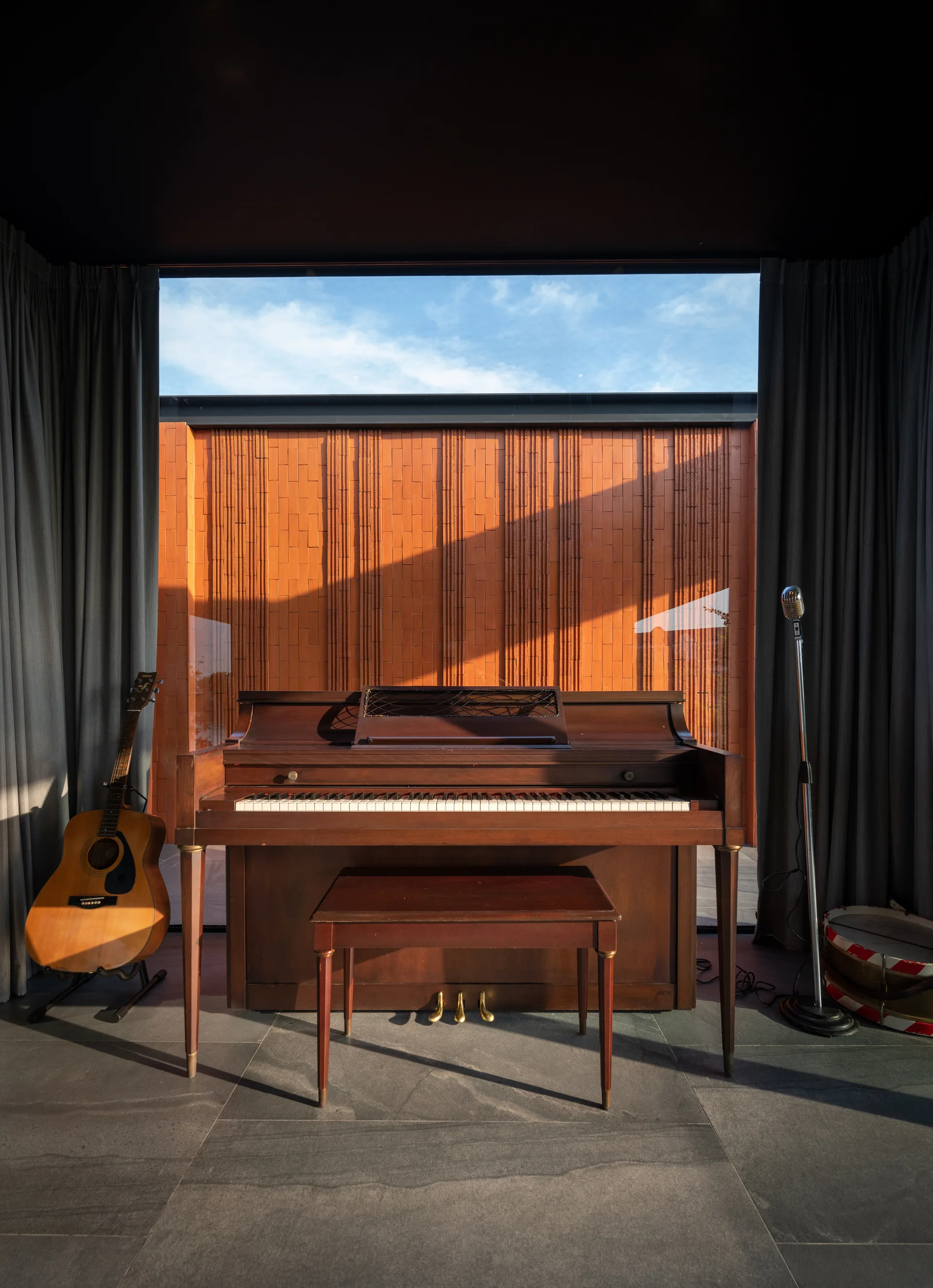 An upright wooden piano and an acoustic guitar stand in front of a large window looking out at a textured terracotta brick wall.