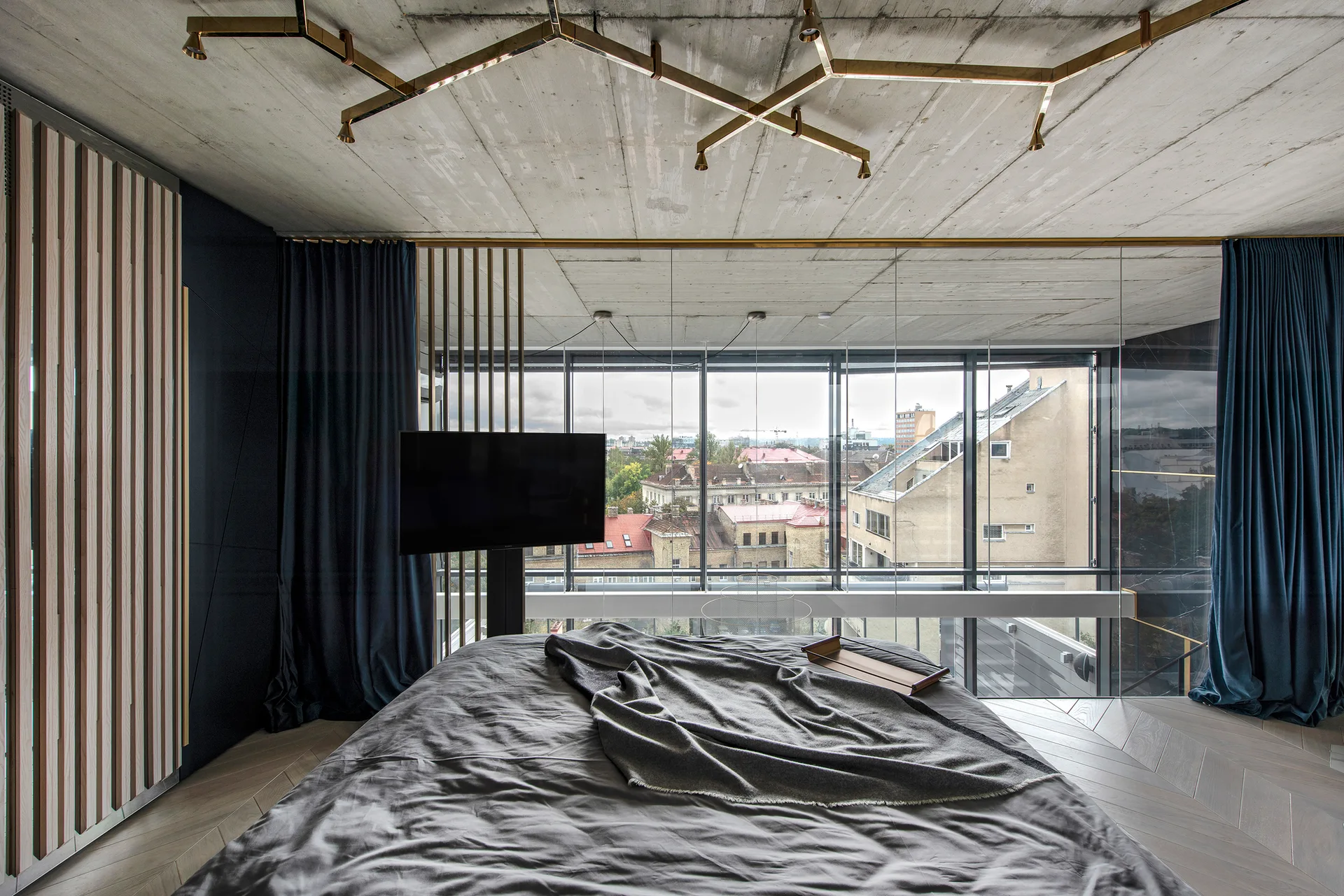 A modern bedroom features an exposed concrete ceiling, a geometric brass light fixture, a bed with grey sheets, and a large window overlooking city buildings.