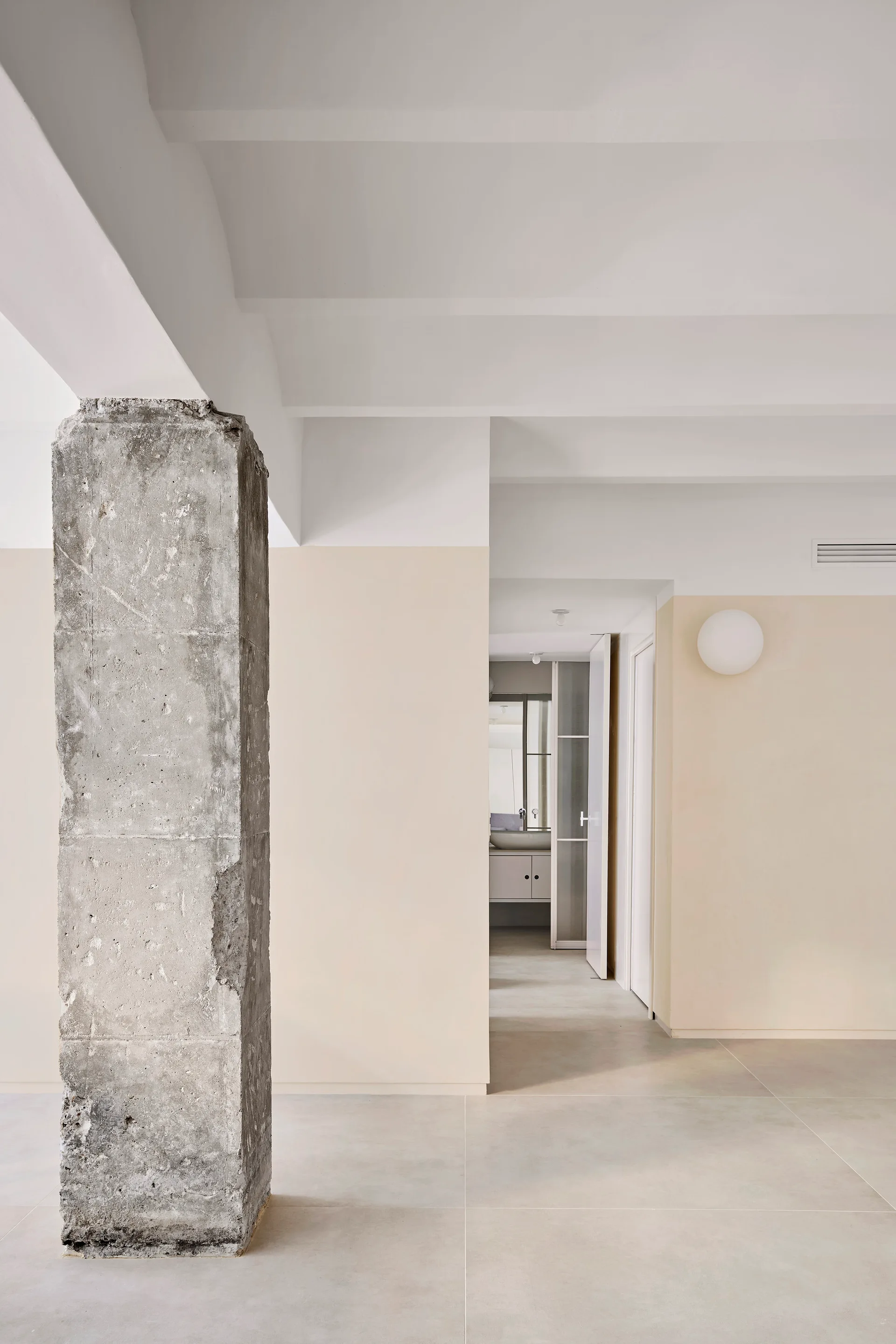 An exposed concrete column stands in a minimalist hallway with cream and white walls, leading to a bathroom doorway with a spherical sconce.