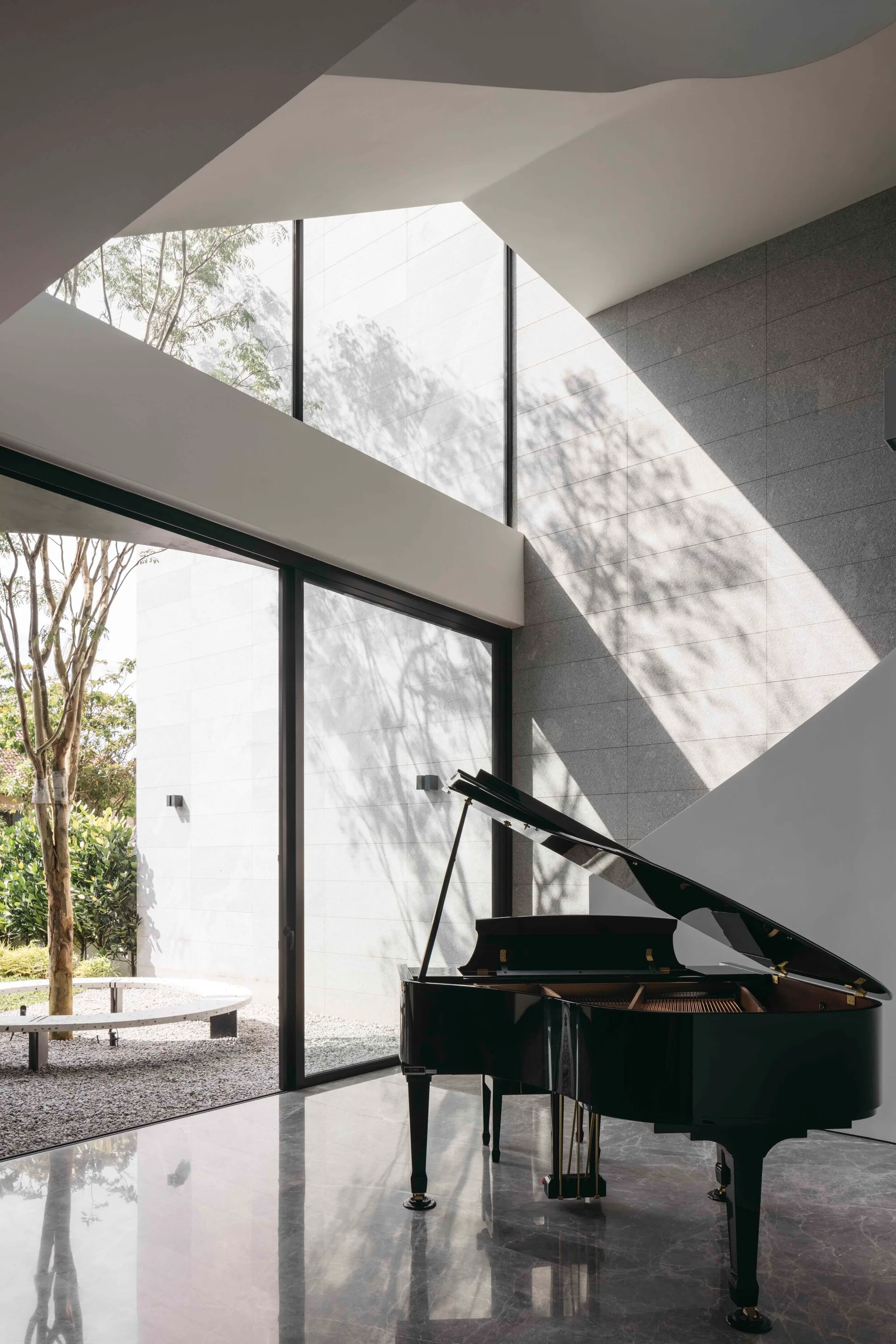 A black grand piano sits on reflective gray marble floors next to floor-to-ceiling glass windows overlooking a gravel courtyard with a tree.