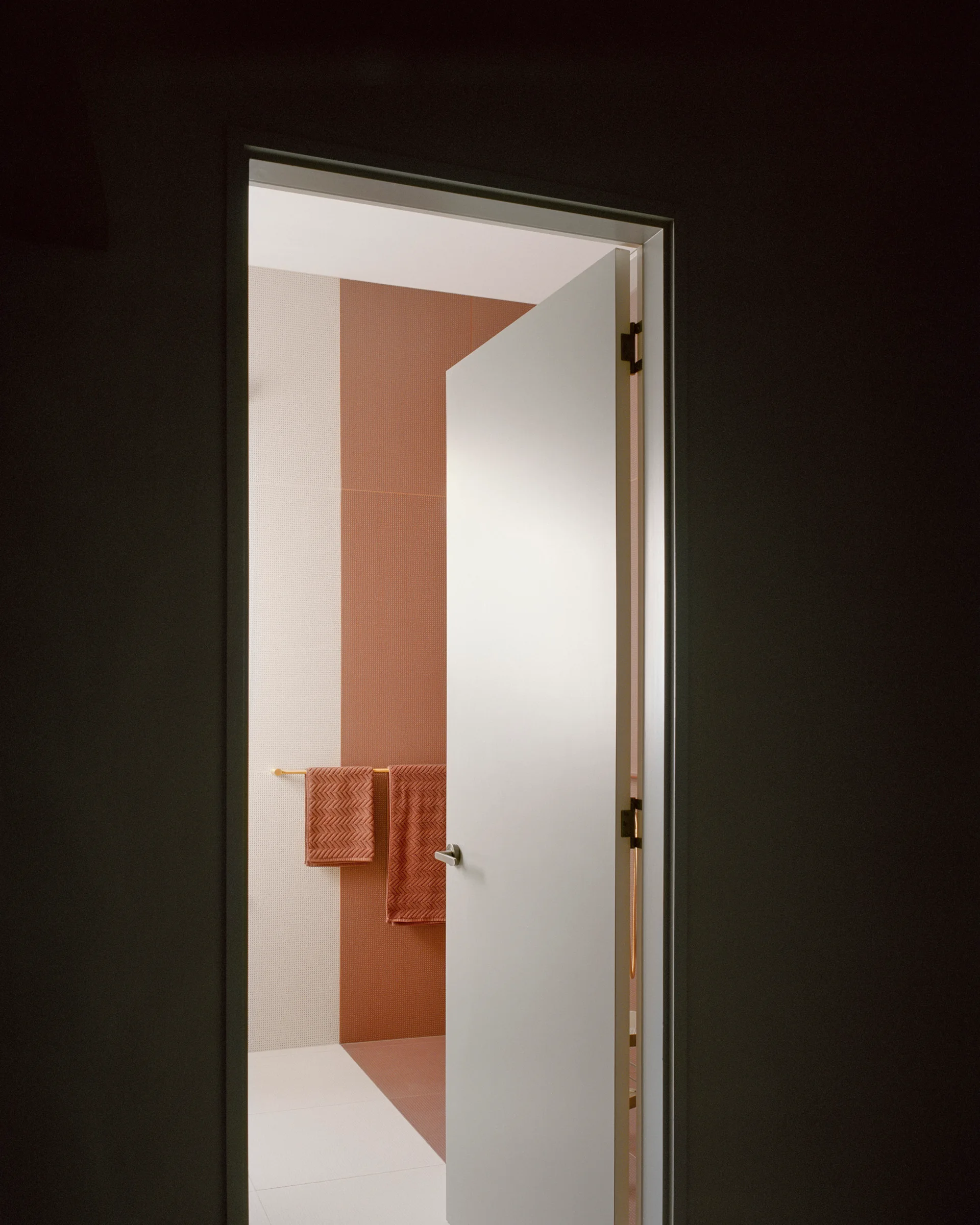 A view through an open white doorway reveals a minimalist bathroom featuring vertical color-blocked wall tiles in off-white and terracotta.