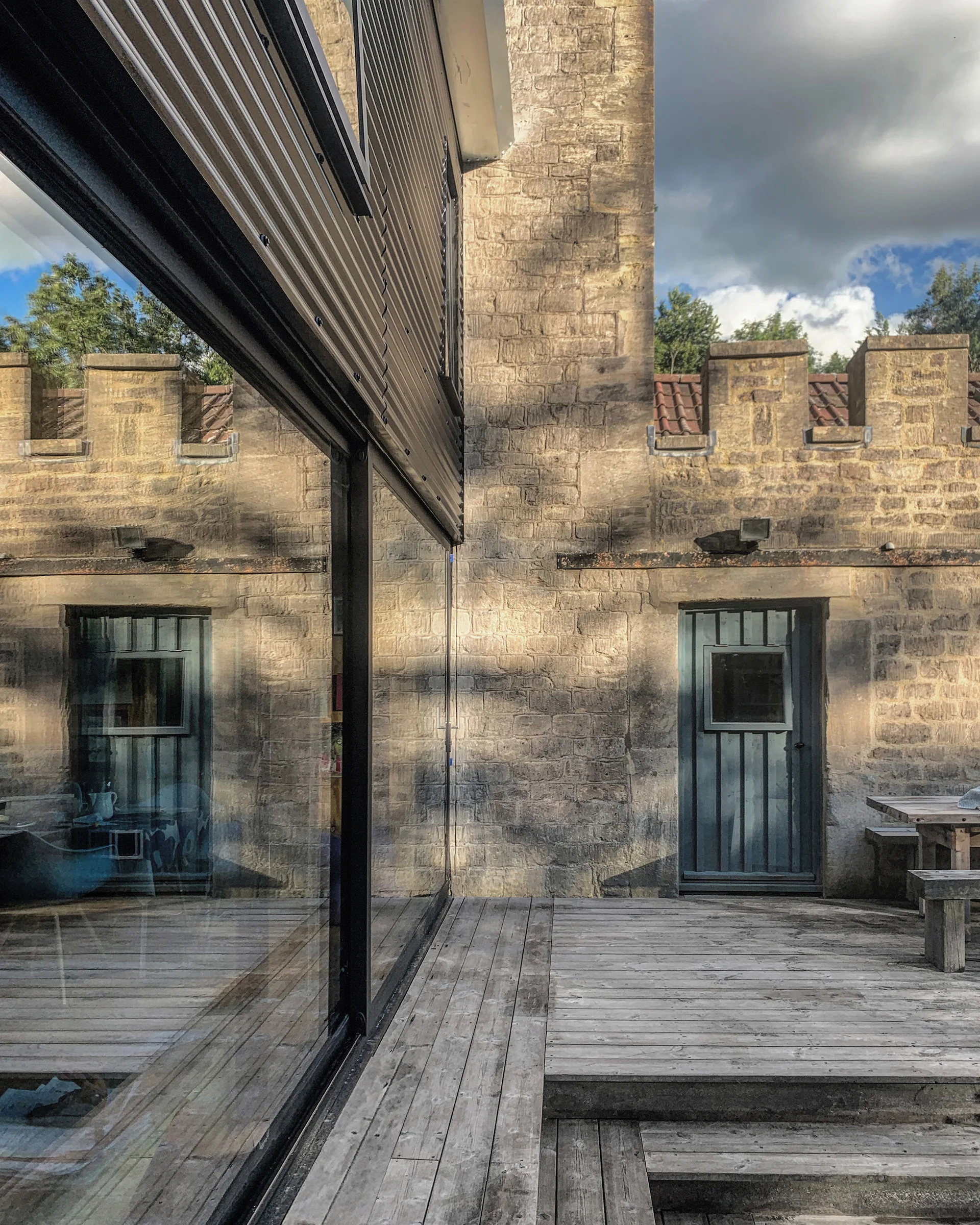 A weathered wooden deck runs alongside a textured stone building featuring a blue-grey door, with a large glass facade reflecting the exterior patio.